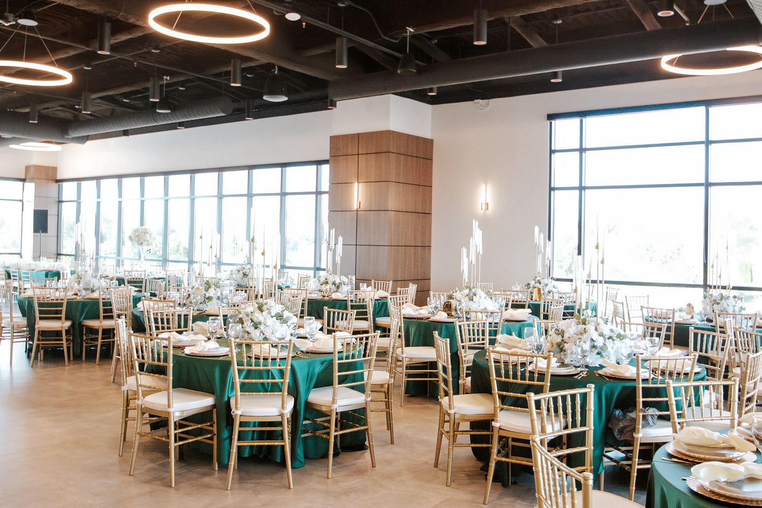 Elegant wedding reception hall with round tables, teal tablecloths, gold chiavari chairs, and circular lighting.