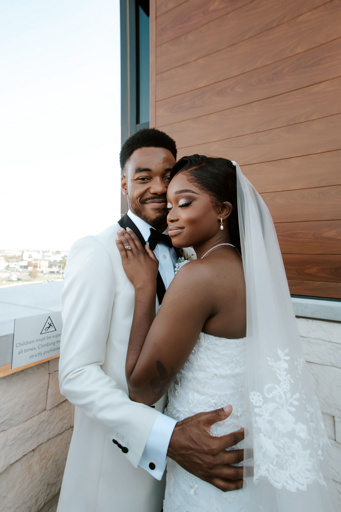 Newlywed couple embracing outdoors, groom in white suit and bride in lace wedding gown with veil.