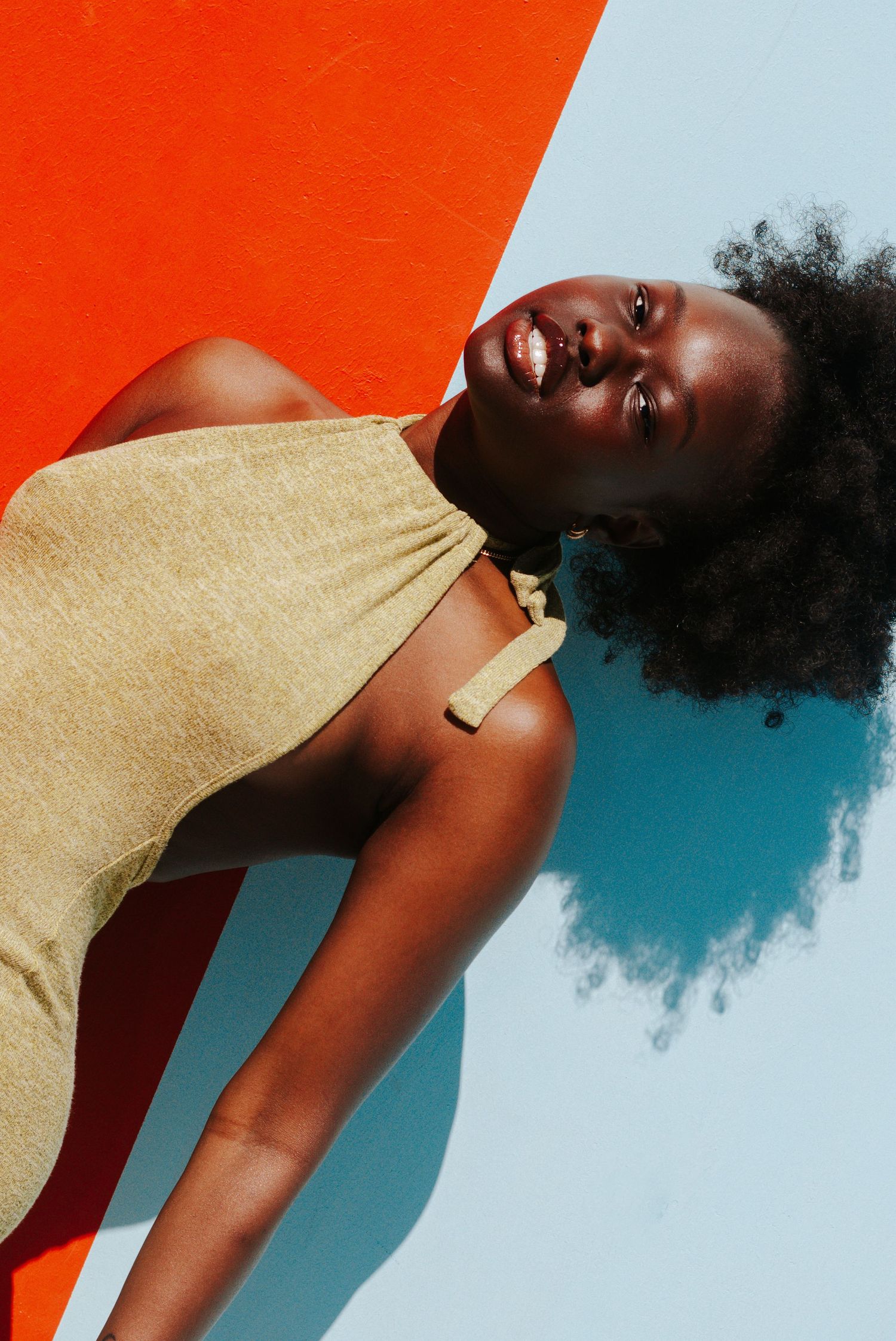 Black woman in gold halter top leaning against bold orange and blue background in sunlight.