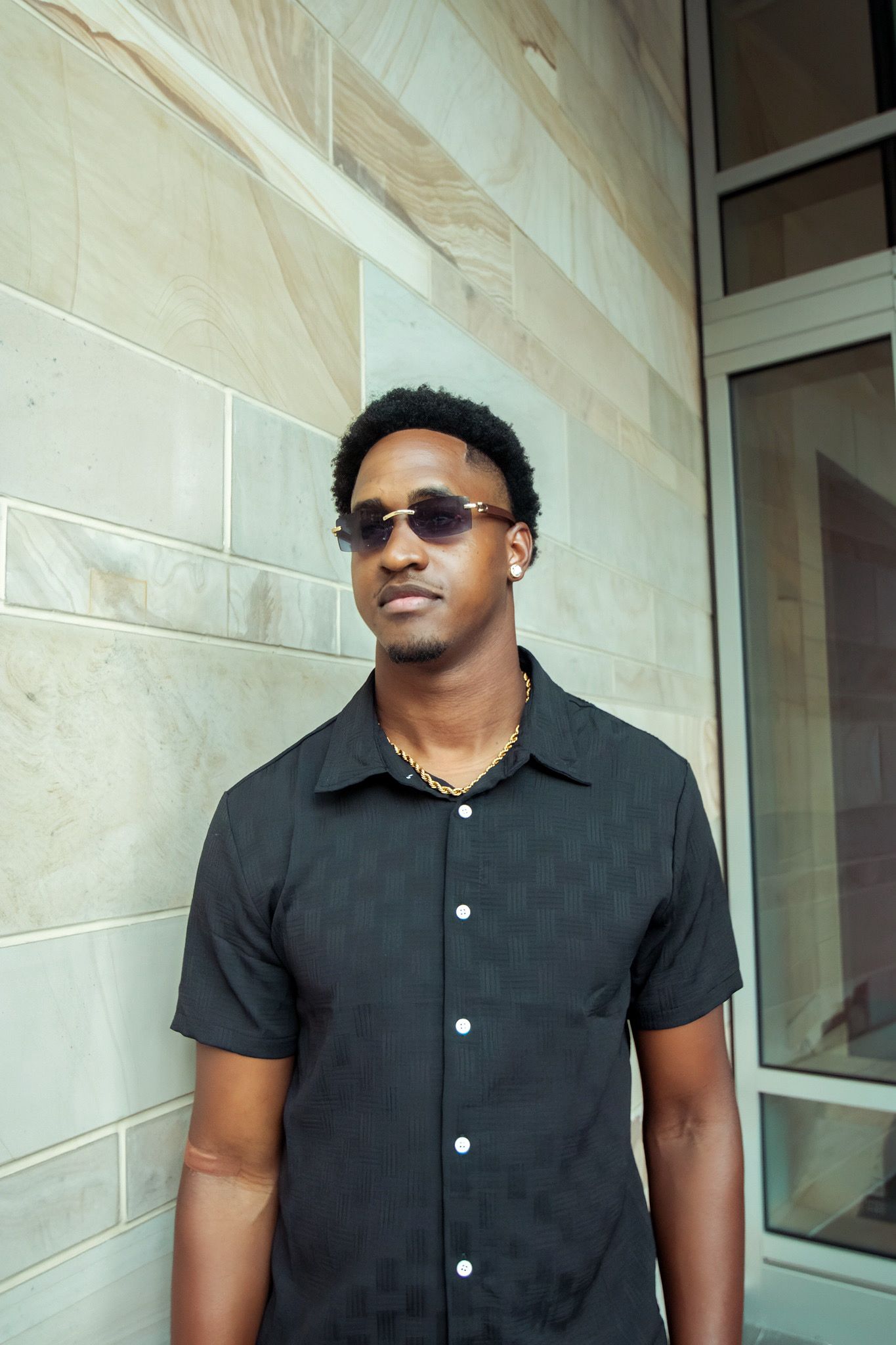Young Black man in black checkered shirt and sunglasses leaning against beige tiled wall outdoors.