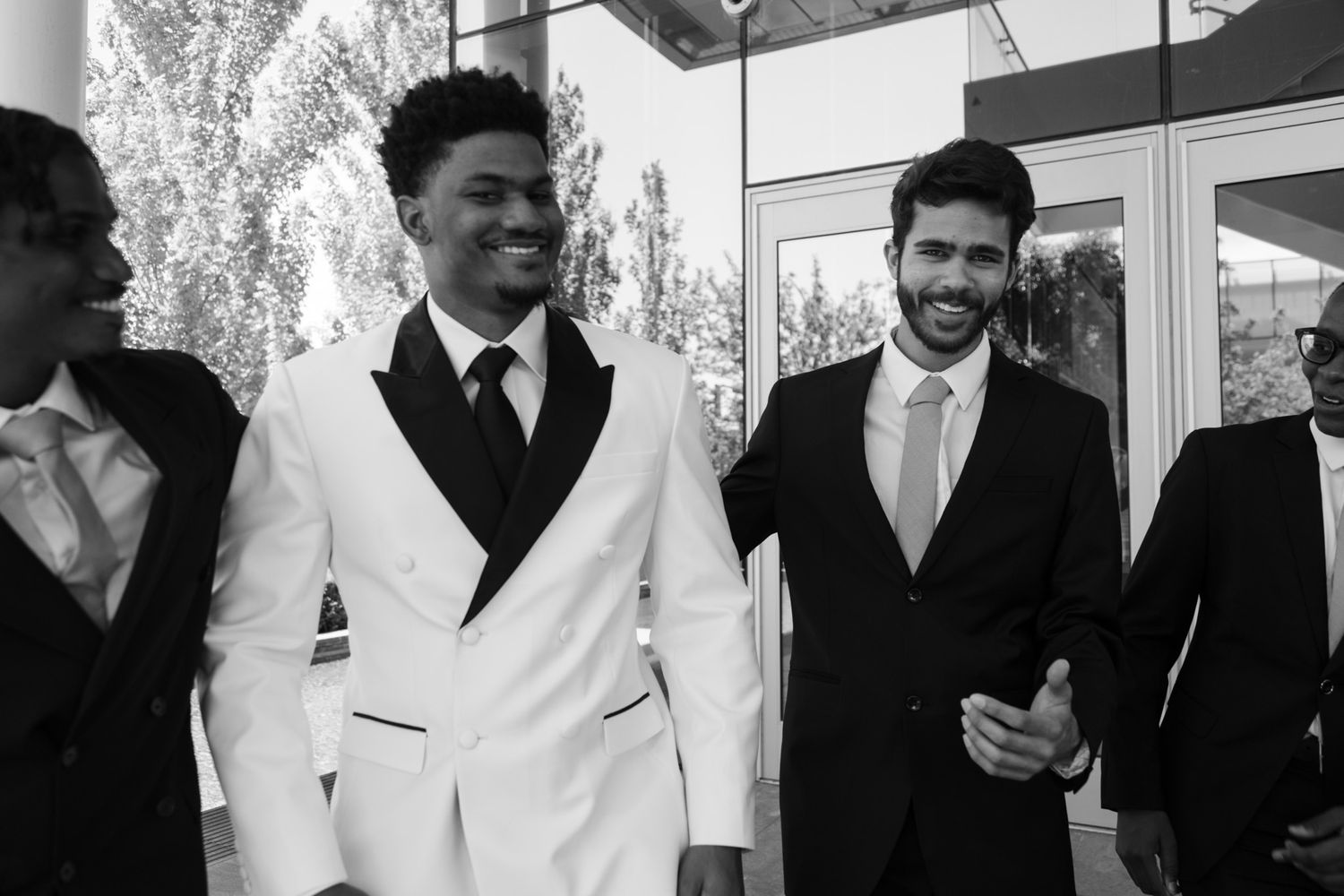 Groom in white tuxedo laughs with groomsmen in black suits outside a modern wedding venue.