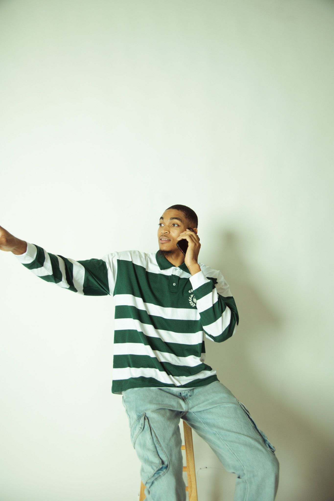 A young man in a striped polo shirt talks on a phone while sitting on a stool in a studio.