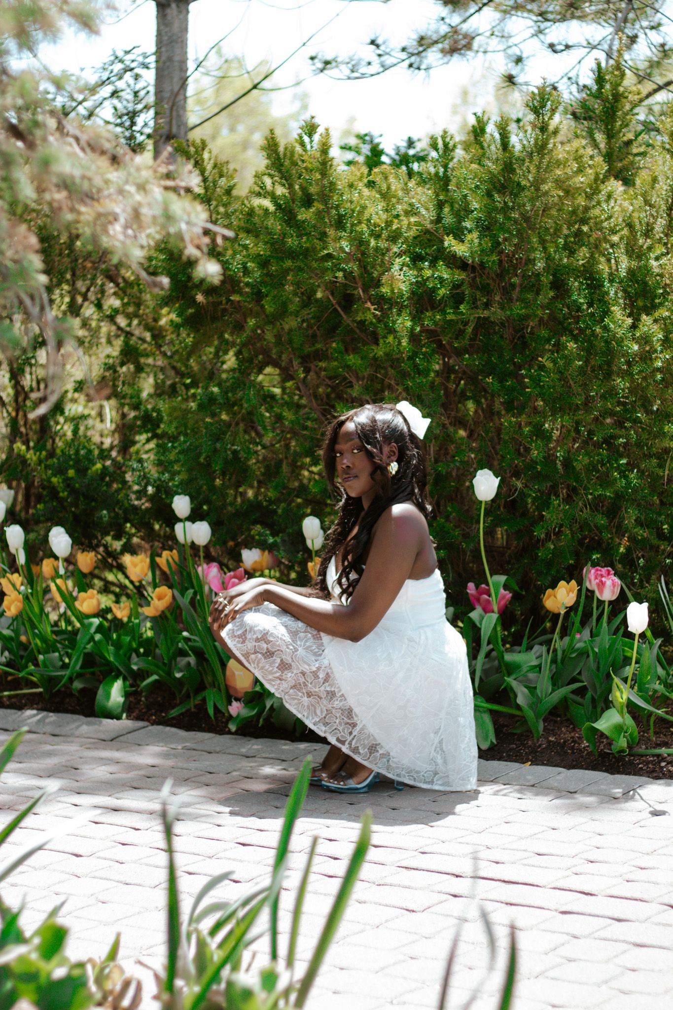 Young girl in white lace dress crouching among colorful tulips in a lush garden.