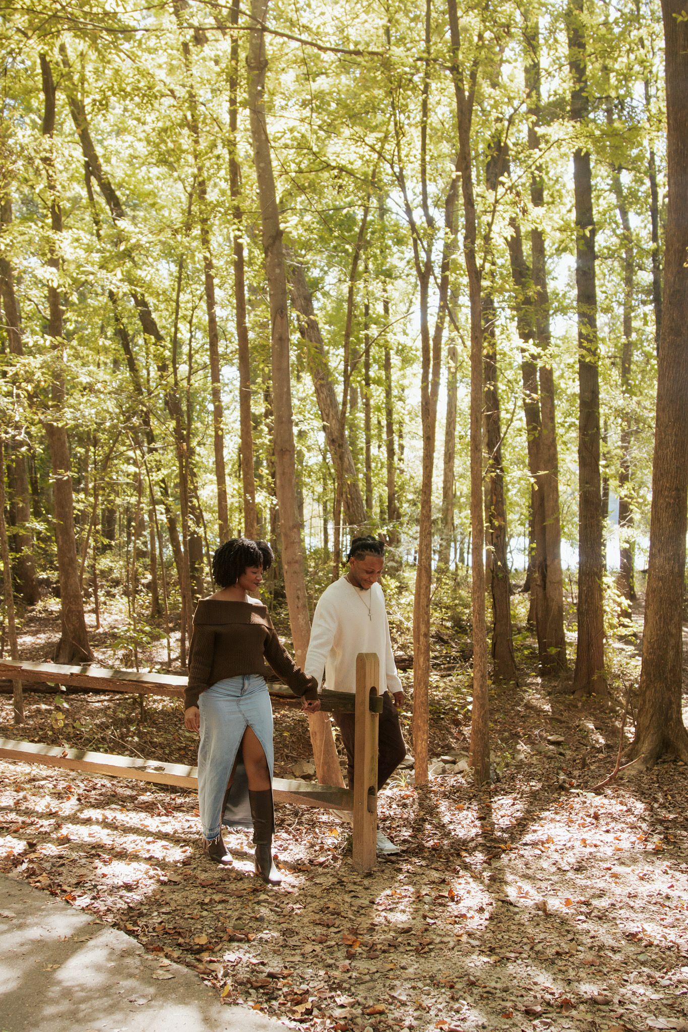Two women sitting on a wooden bench in a sunlit forest with tall trees and fallen leaves.