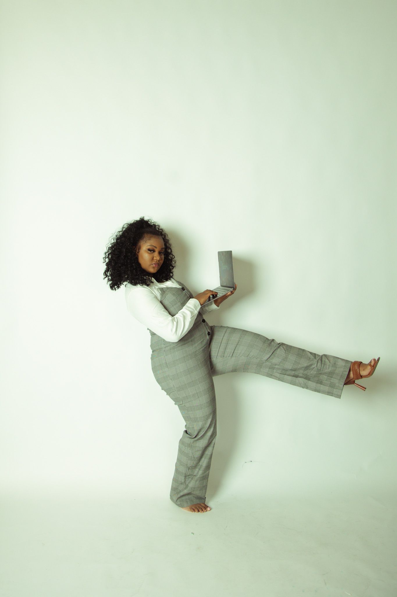A woman in gray overalls and white top poses dynamically with a laptop in a studio.