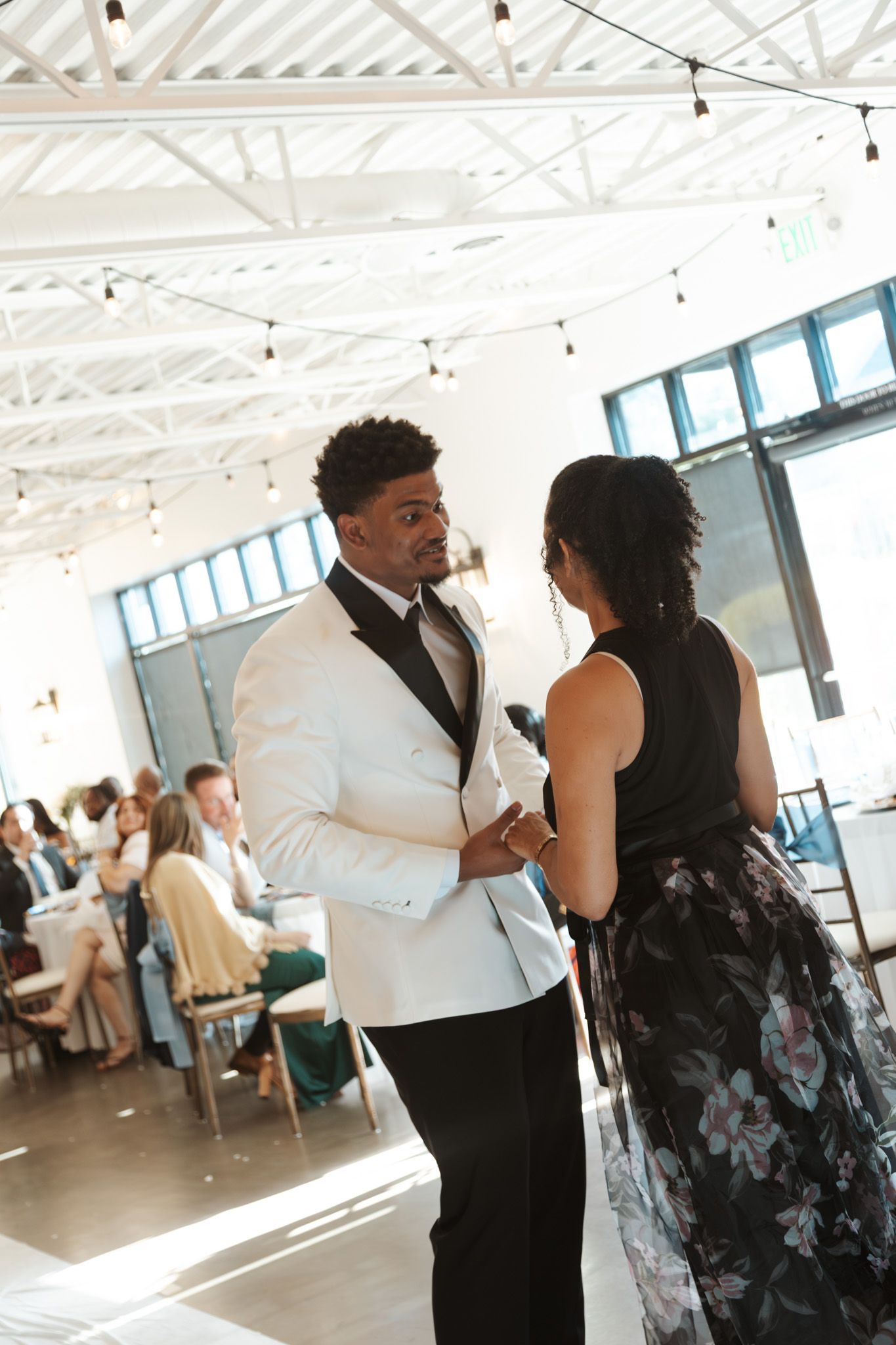 Groom in white tuxedo shares a dance with a woman in black floral dress at wedding reception.