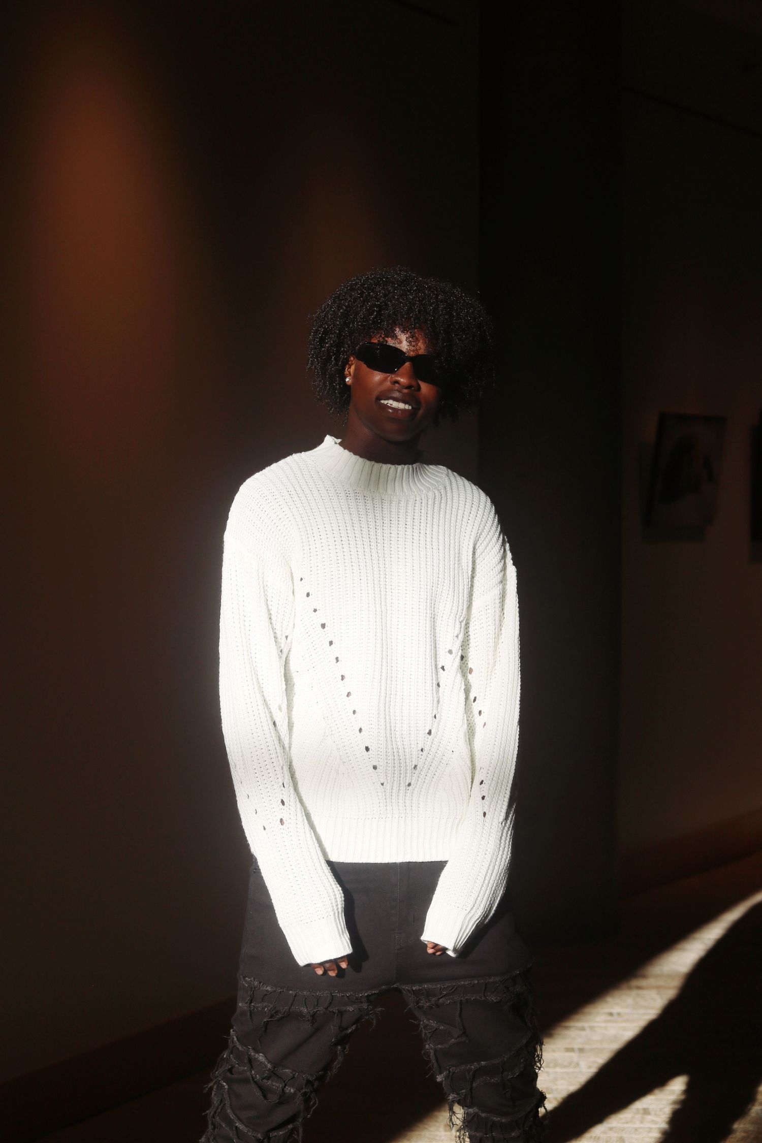 Young Black man smiling in white ribbed knit sweater and dark sunglasses, standing in dramatic sunlight.