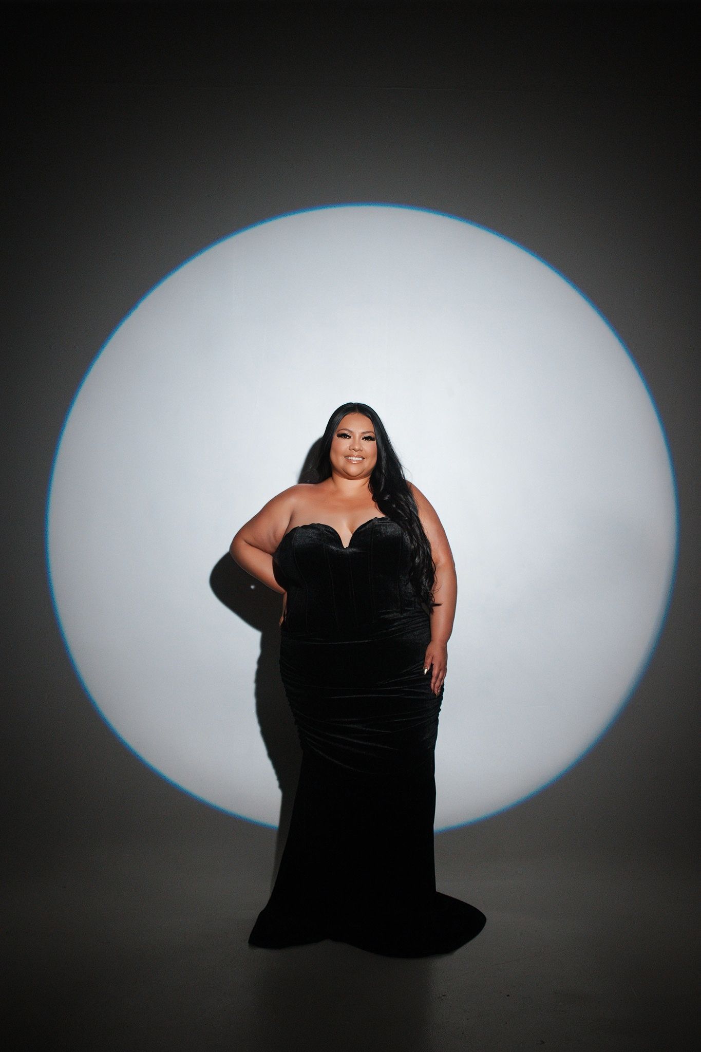 Plus-size woman in elegant black strapless velvet gown posing in front of a circular studio light.