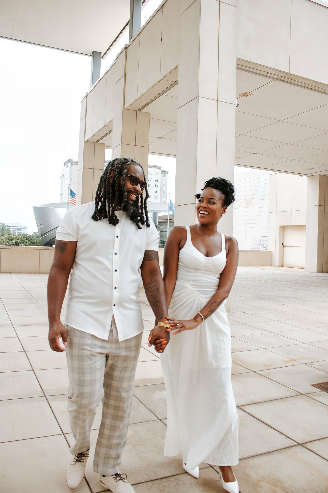 Smiling couple in matching white outfits holding hands outside a modern building.