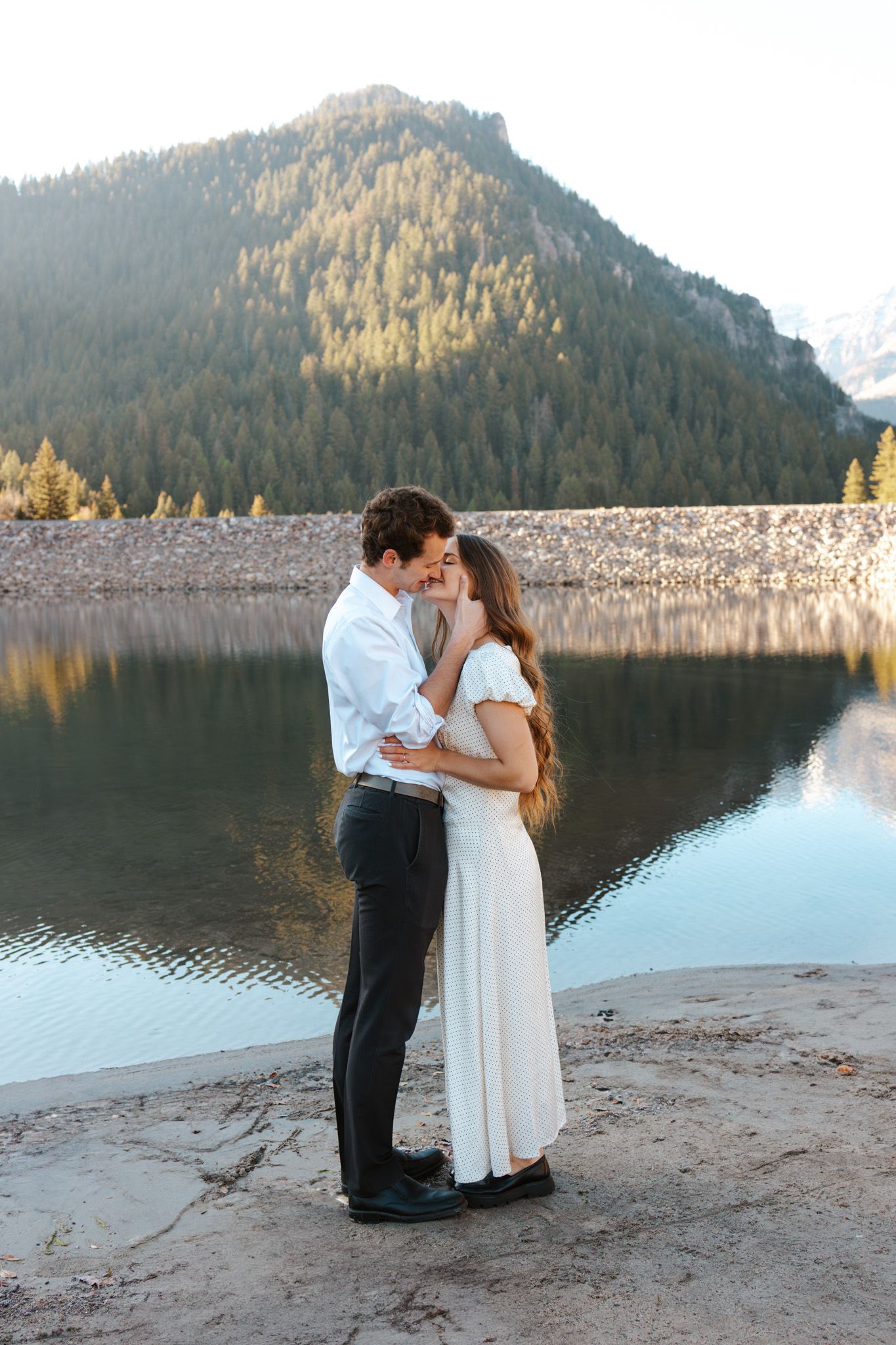 Couple sharing a romantic kiss by a mountain lake at golden hour, woman in white dress.