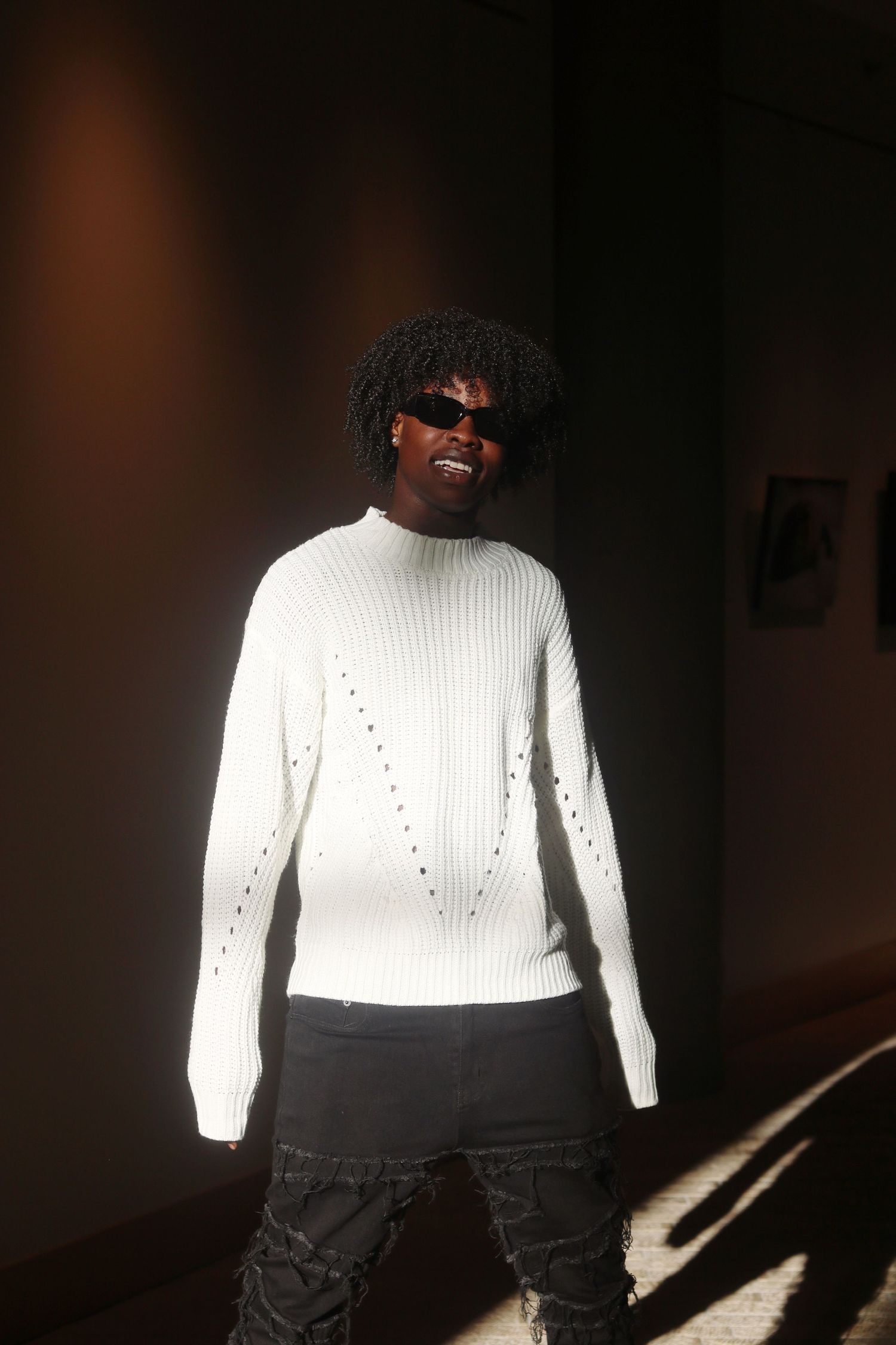 Stylish Black man in white distressed ribbed sweater and sunglasses posing in sunlit indoor space.