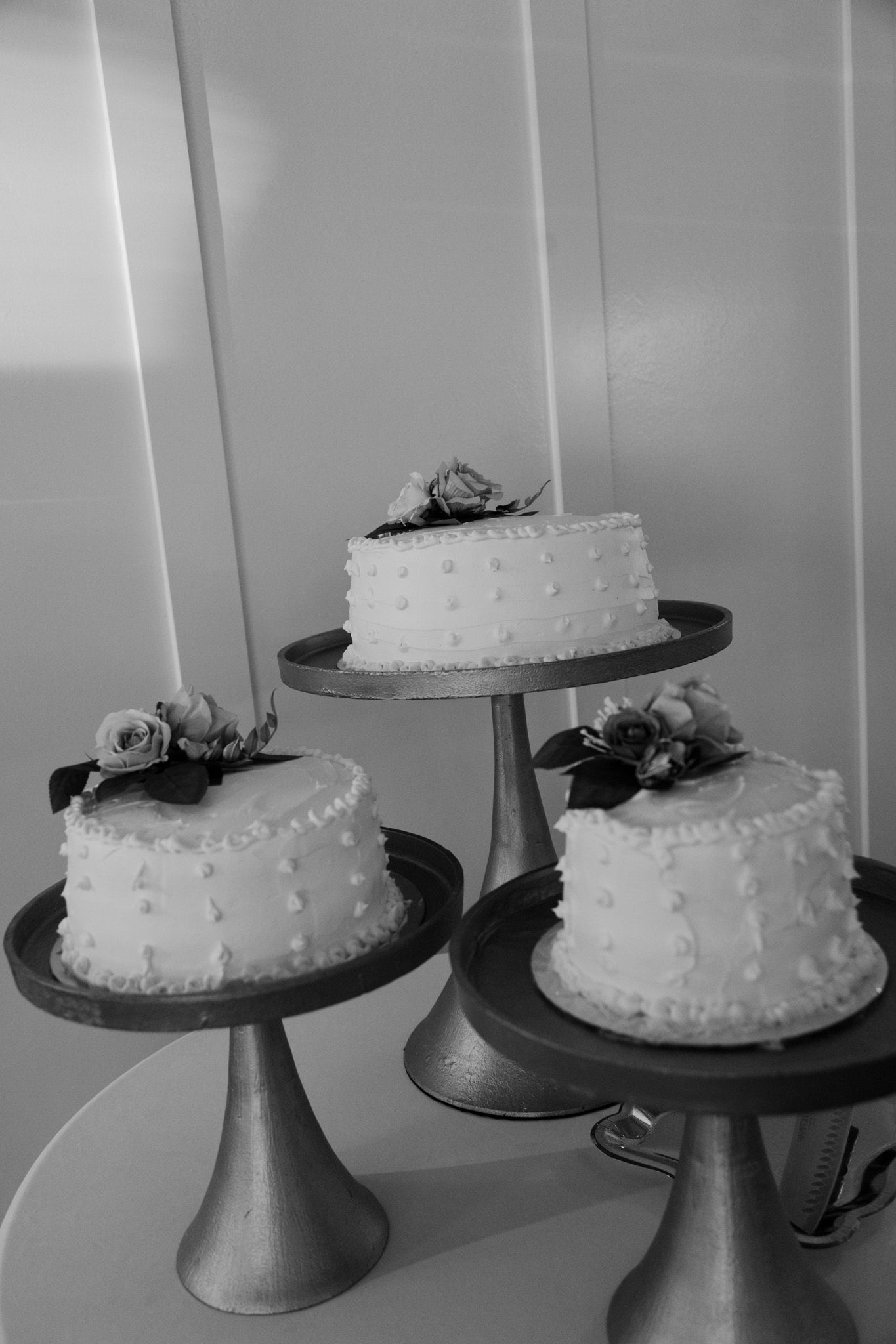 Three elegant white wedding cakes with rose decorations displayed on gold stands in black and white.