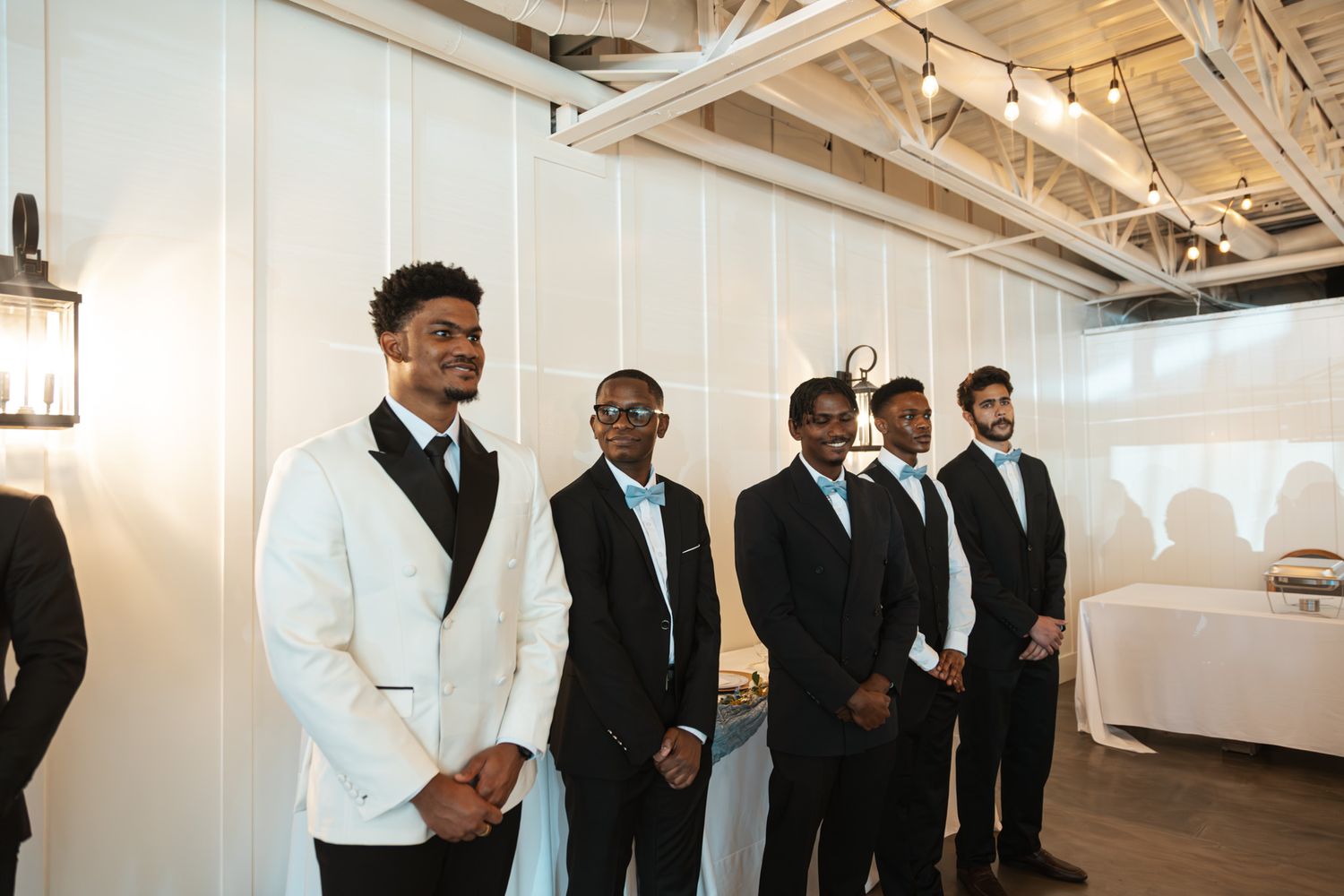 Groomsmen in black tuxedos stand beside groom in white jacket at indoor wedding venue.