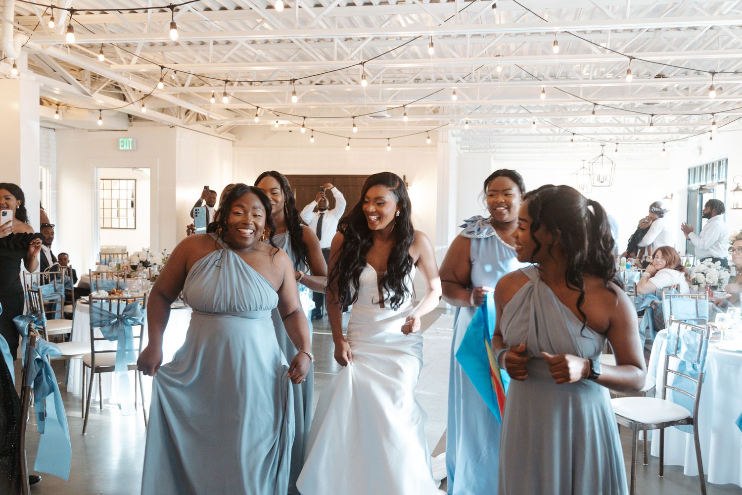 Bride in white gown walks joyfully with bridesmaids in light blue dresses at wedding reception.