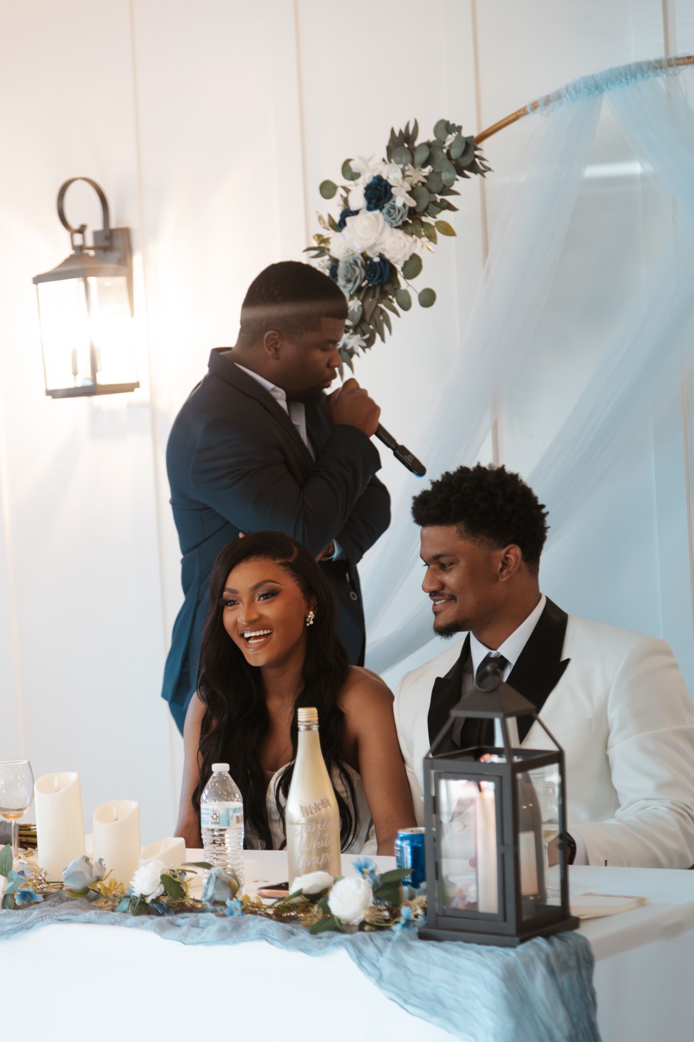 Best man gives toast holding bouquet beside smiling bride and groom at reception table.