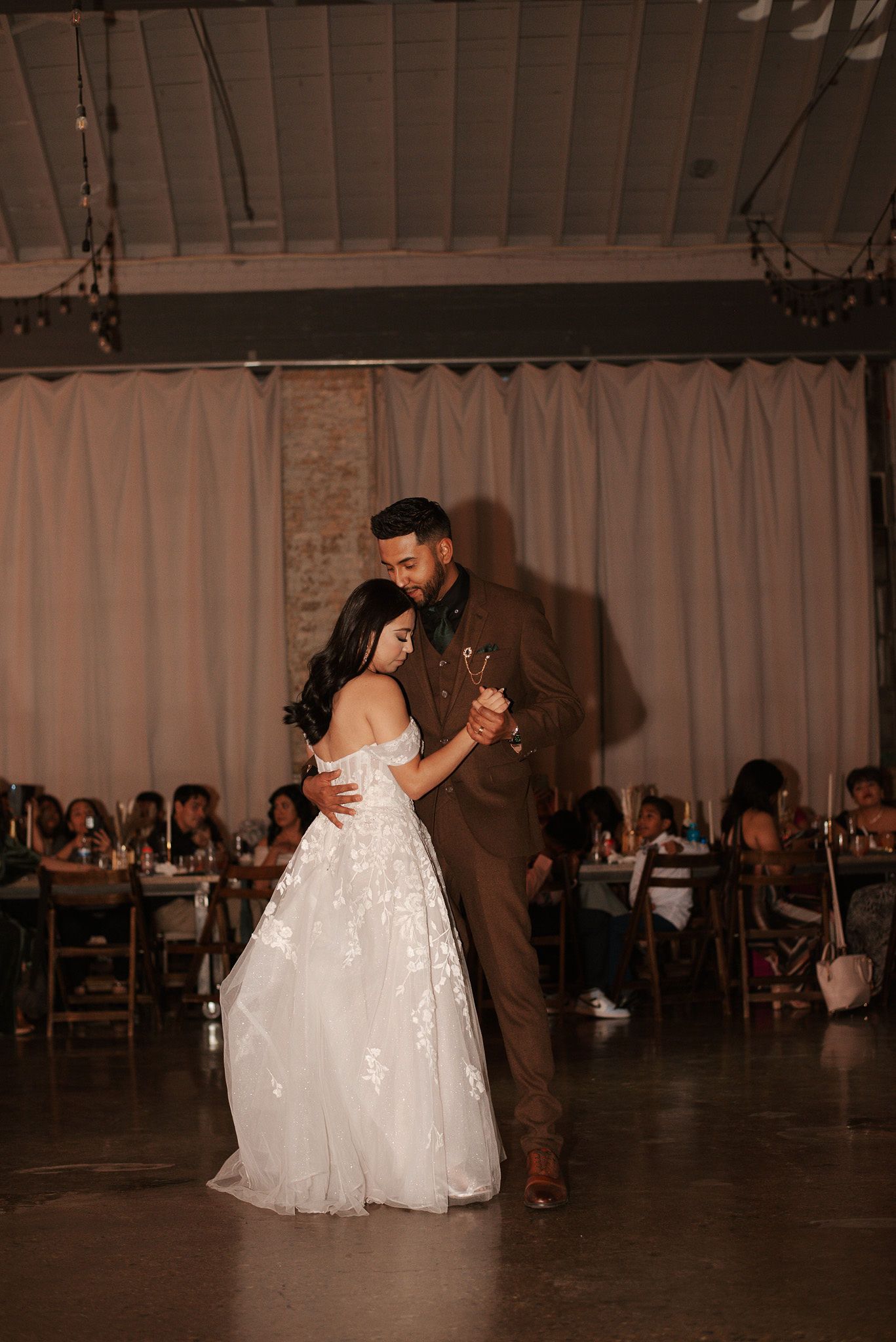 Bride in off-shoulder gown and groom in brown suit share their first dance at wedding reception.