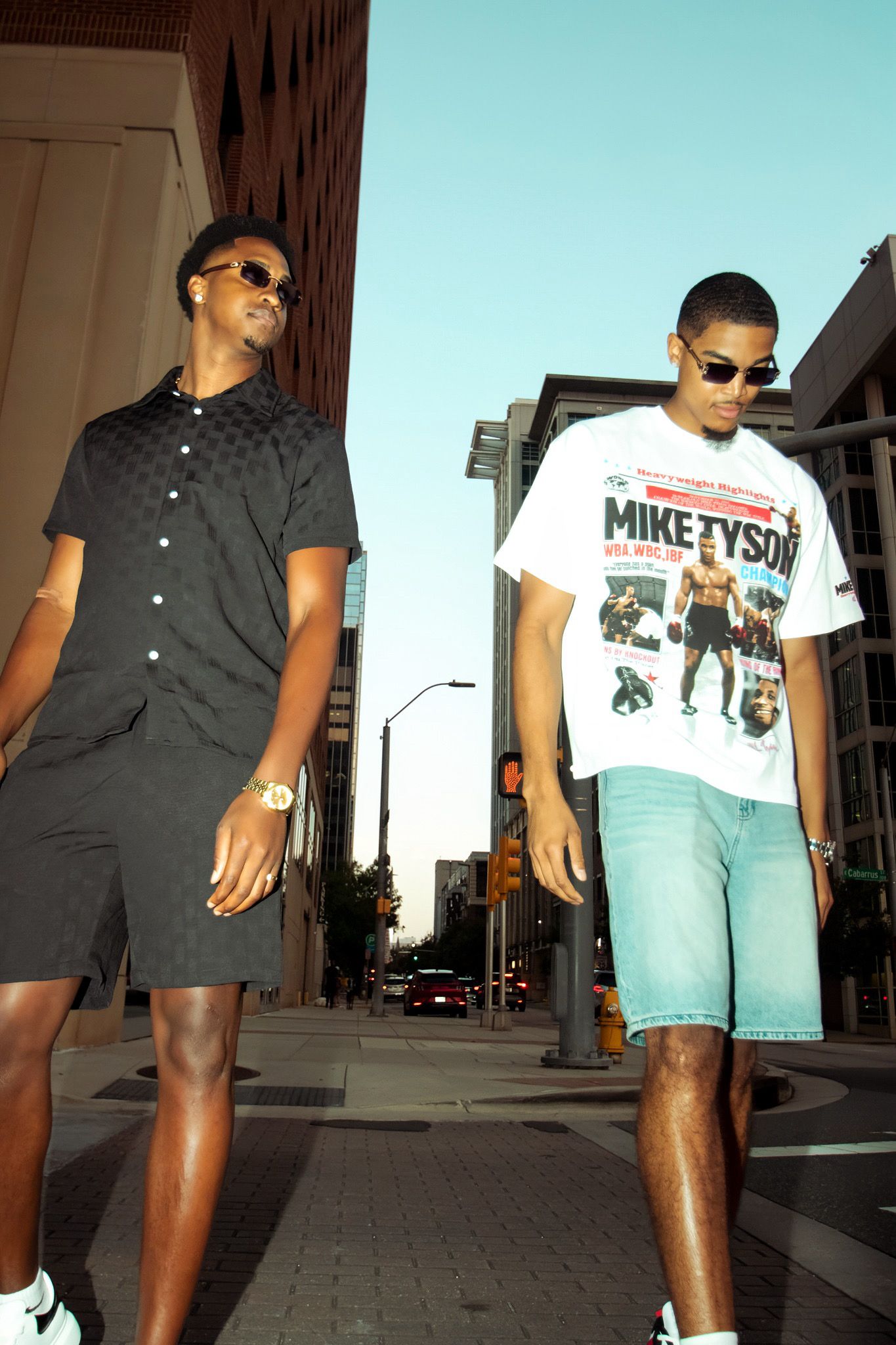 Two stylish young Black men in sunglasses walking on city street in coordinated casual outfits.