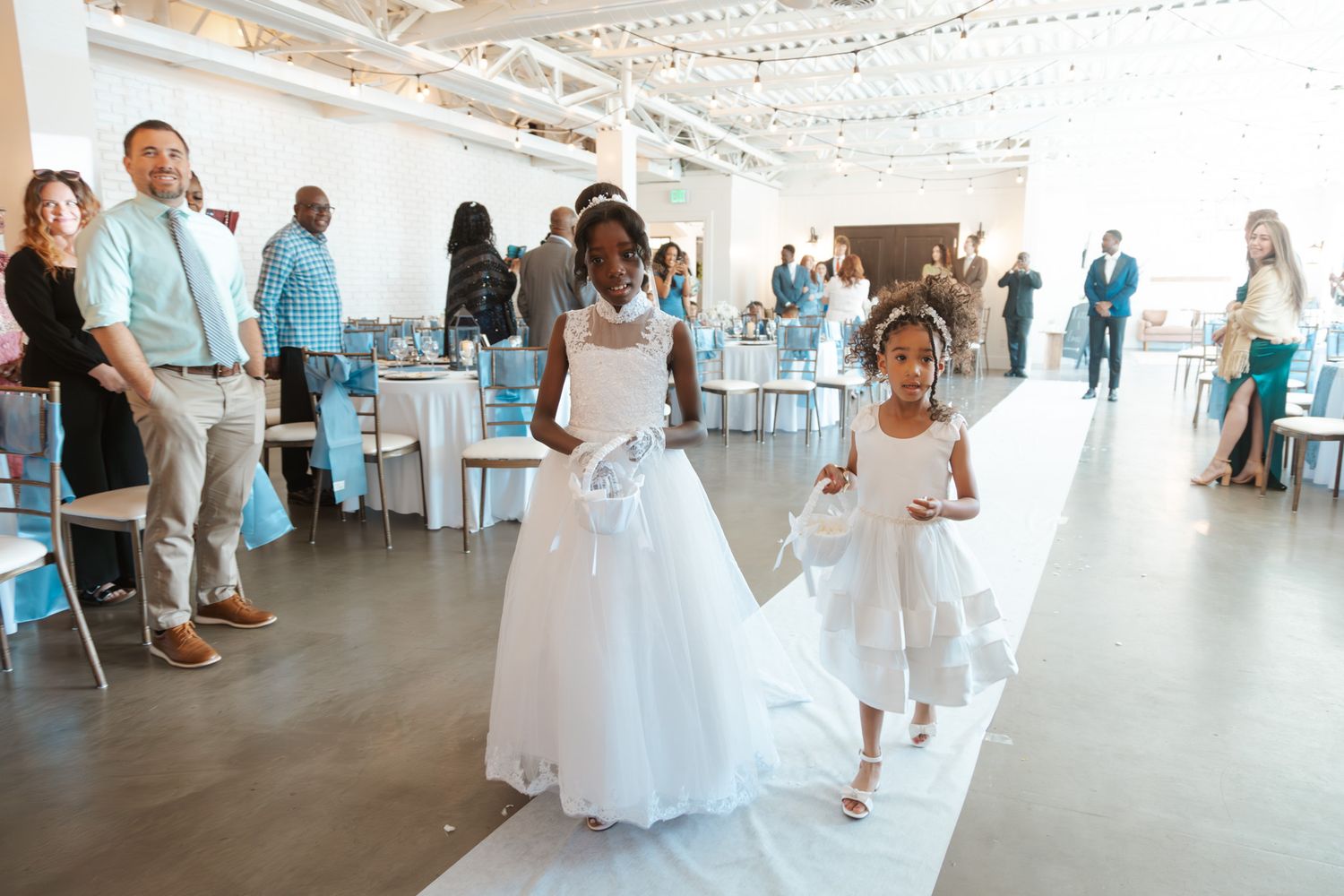 Young flower girls walk down the aisle in white dresses at an elegant wedding reception venue.
