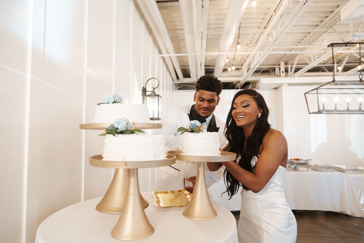 Bride and groom smile while cutting tiered white wedding cake at bright indoor reception venue.