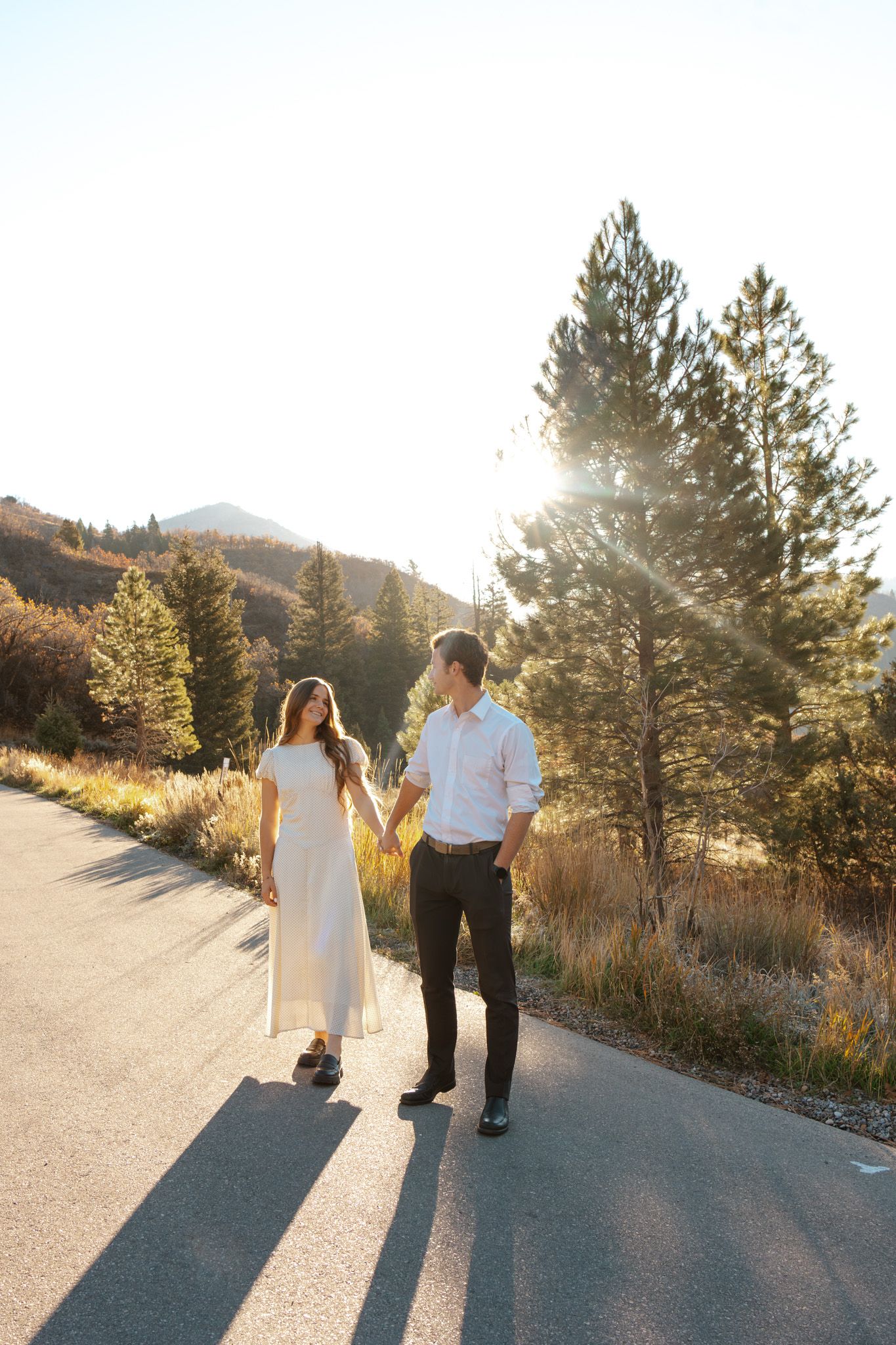 Couple walking hand-in-hand on a sunlit mountain road surrounded by pine trees.