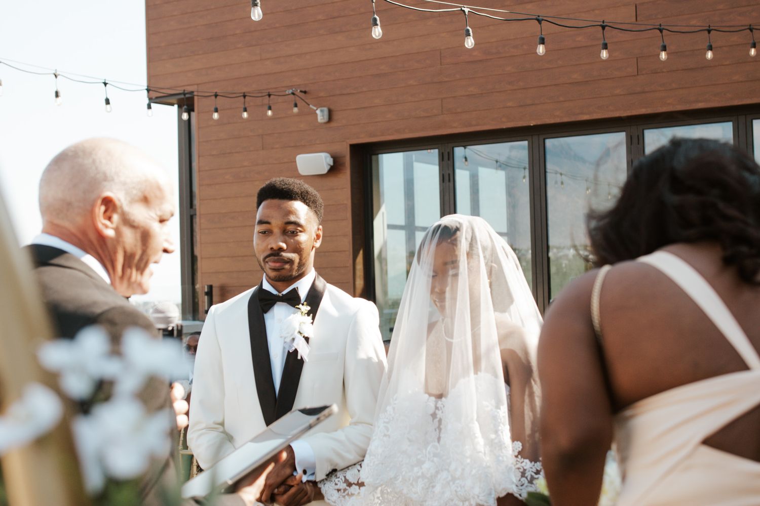 Groom in white tuxedo smiling at veiled bride during intimate outdoor wedding ceremony with officiant.