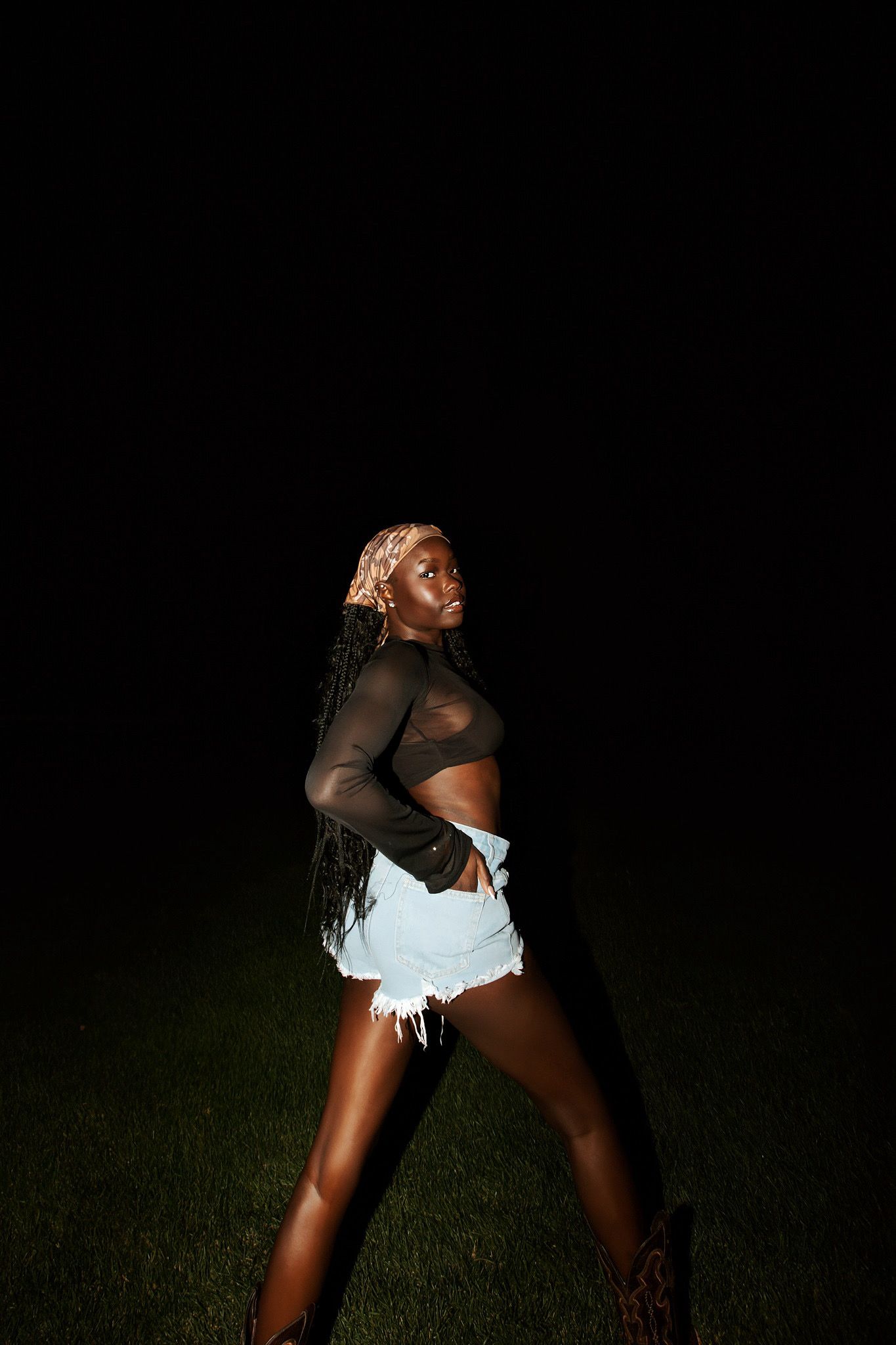 Black woman in denim shorts and sheer top posing on grass against a dark night sky.