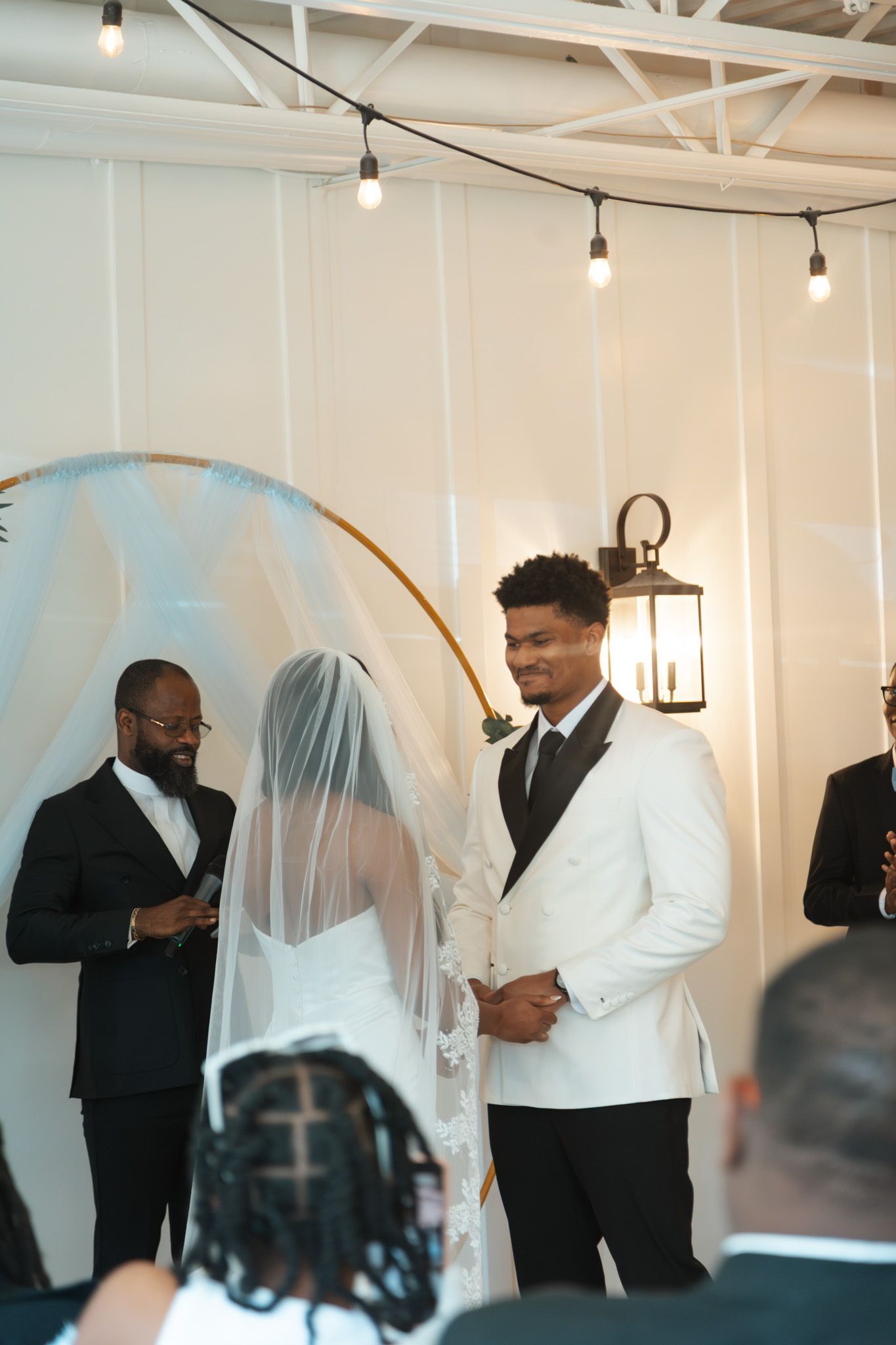 Groom in white tuxedo smiles at bride during indoor wedding ceremony with golden arch.