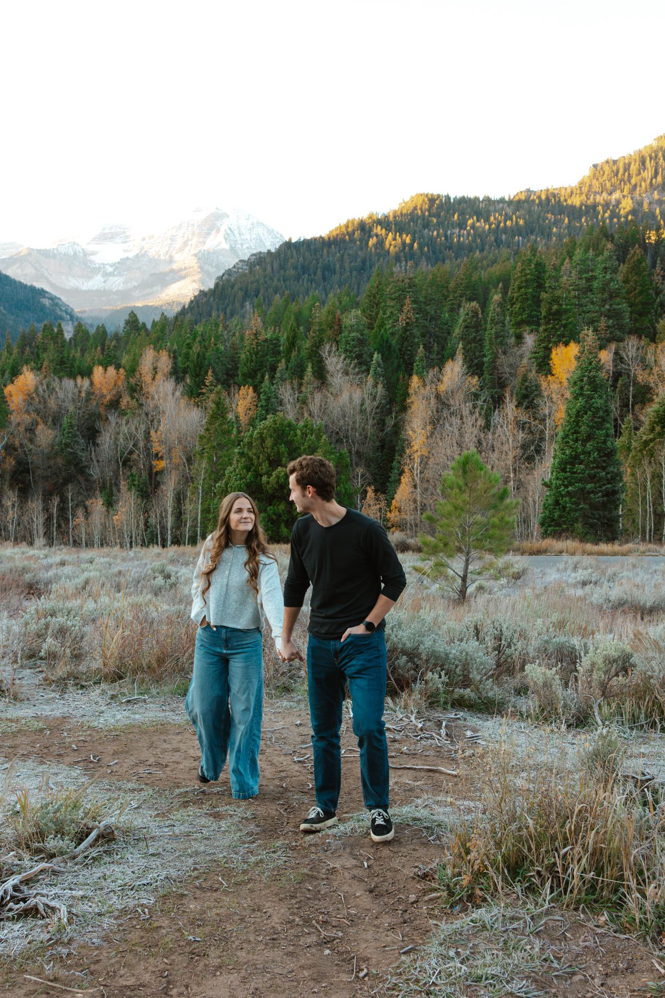 Couple holding hands walking on a dirt path with autumn mountain forest backdrop.