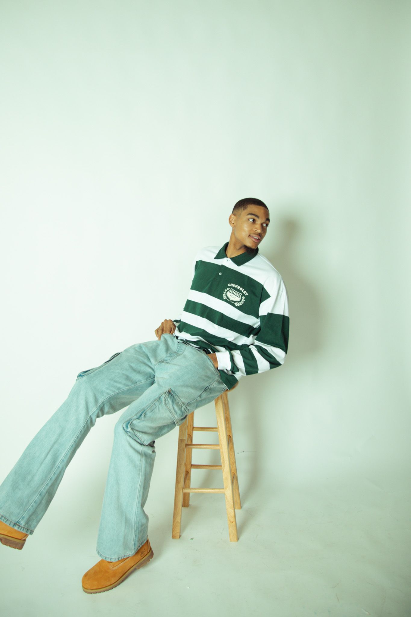 A young man in a green striped shirt and wide-leg jeans sits relaxed on a wooden stool.