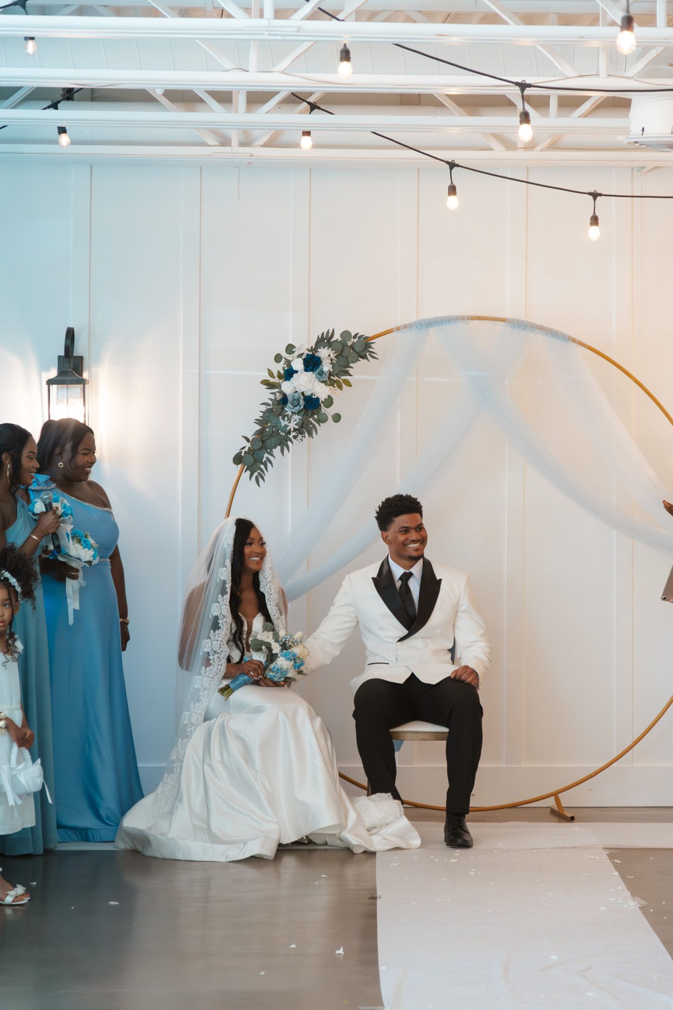 Bride and groom sit together at altar beneath floral hoop arch with bridesmaids in blue.