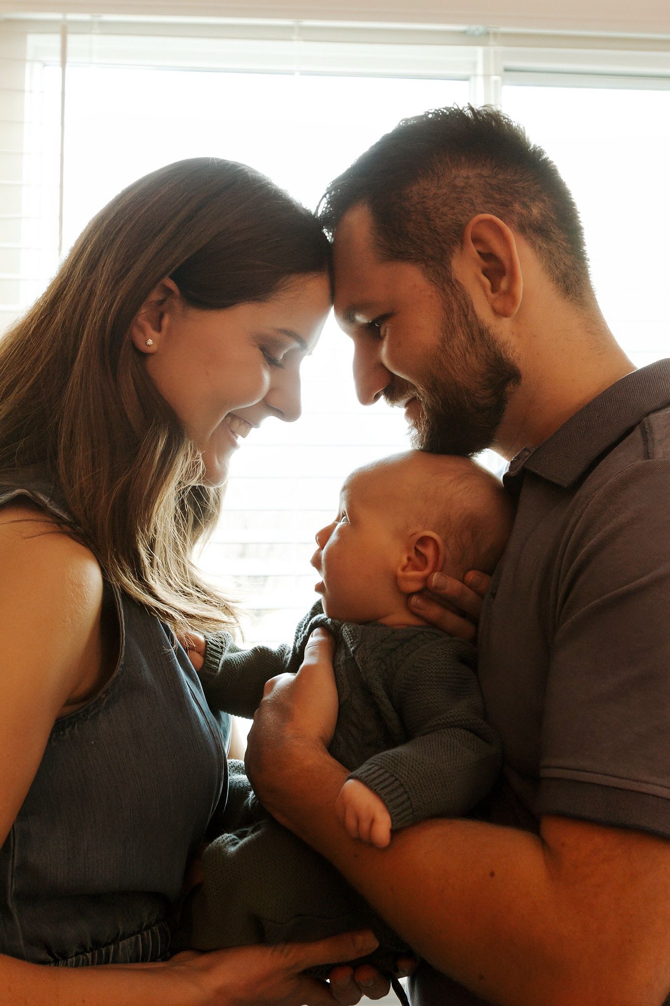 Happy parents smiling and touching foreheads while lovingly cradling their newborn baby together near a bright window.