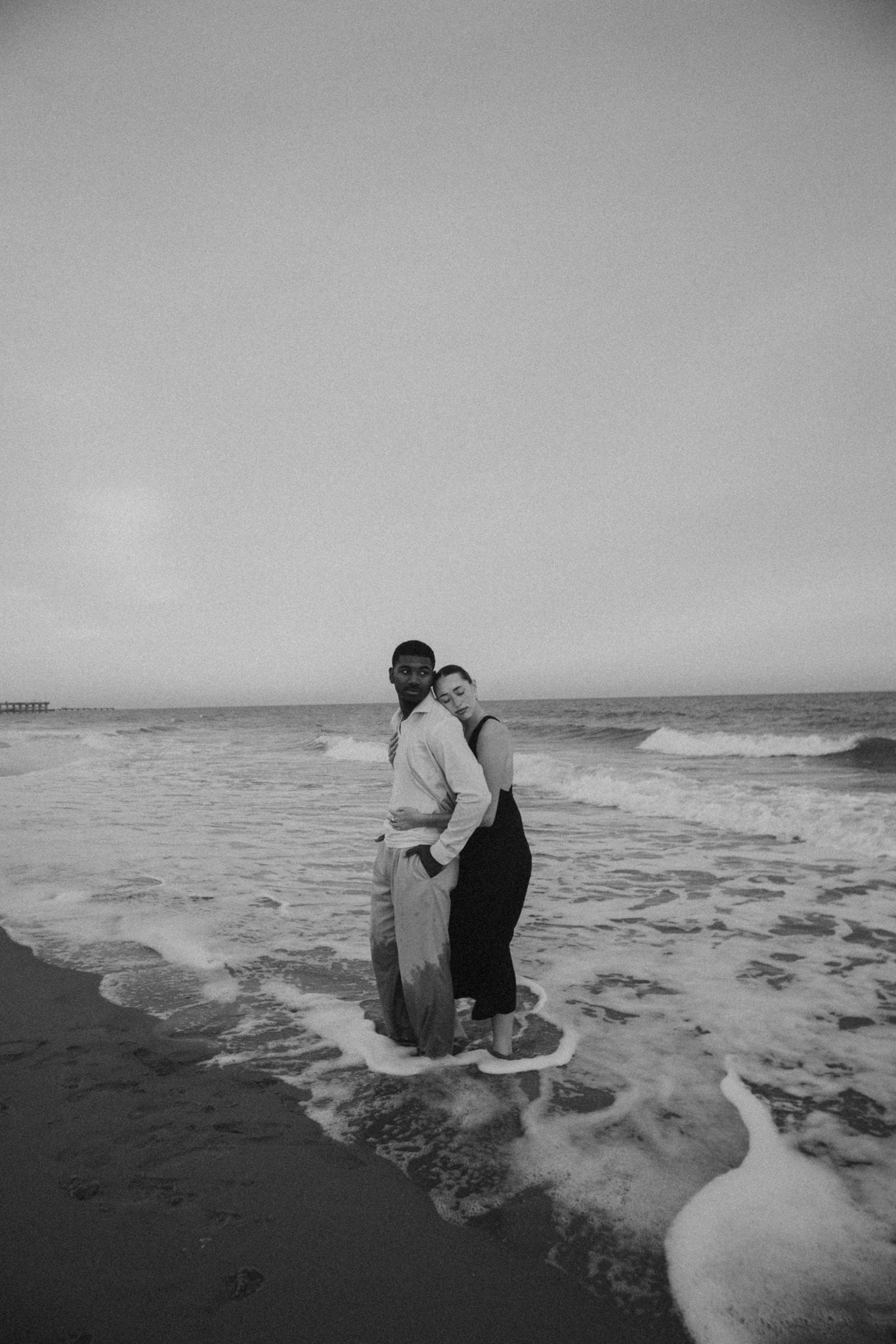 Black and white photo of a couple standing in ocean waves on a beach at dusk.