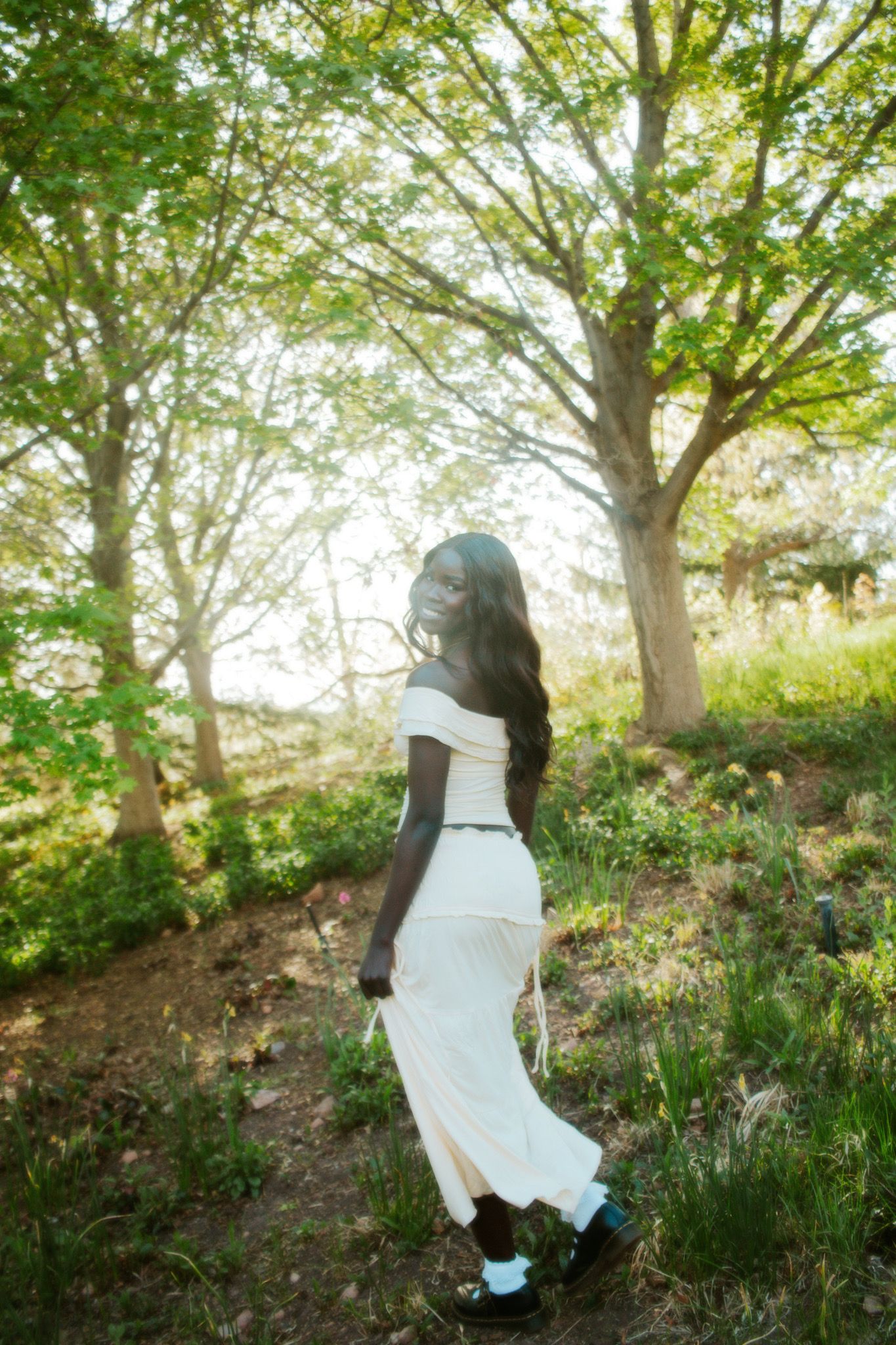 Woman in white off-shoulder midi dress walking through a sunlit green park with trees.