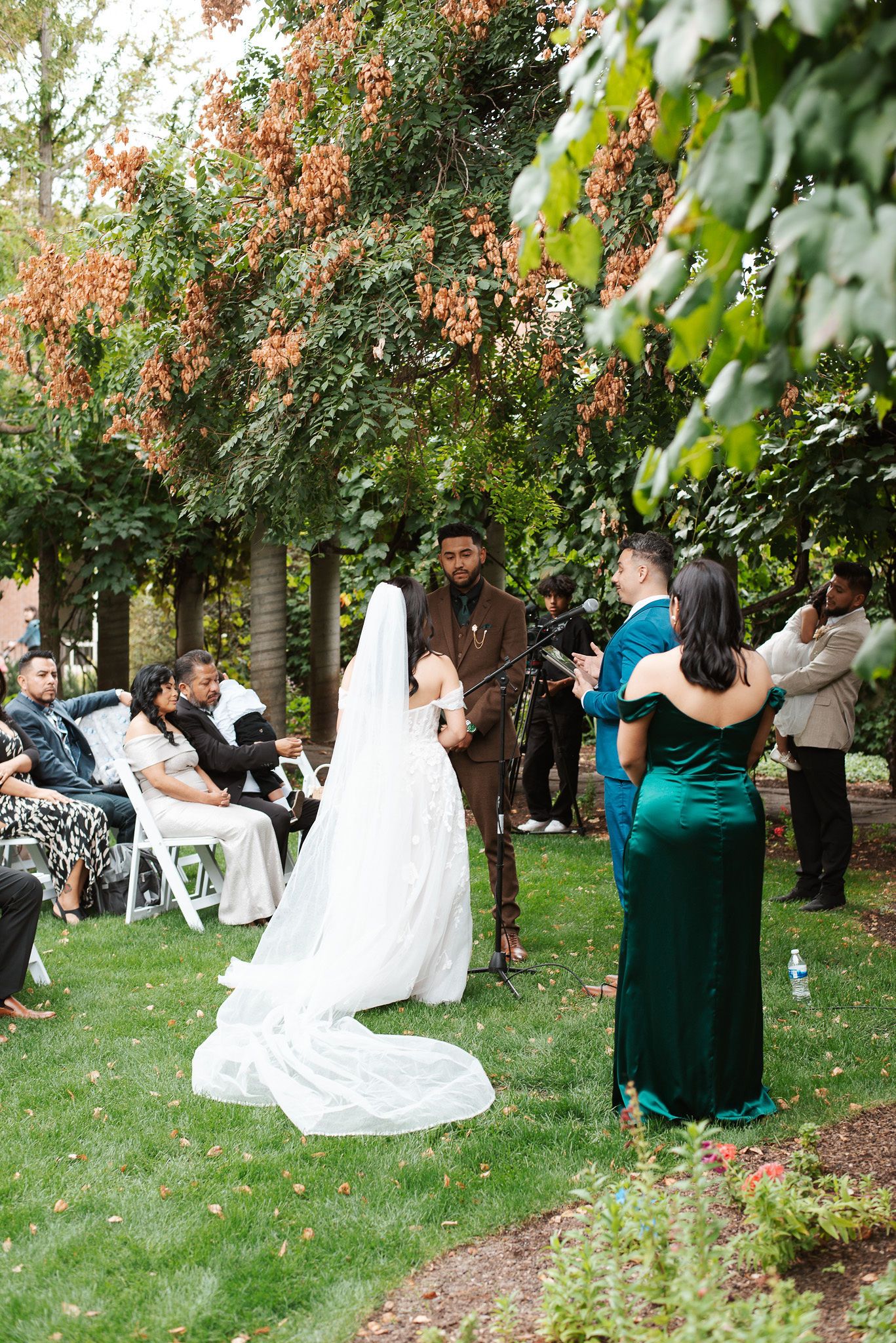 Bride in cathedral veil exchanges vows with groom in brown suit at garden ceremony.