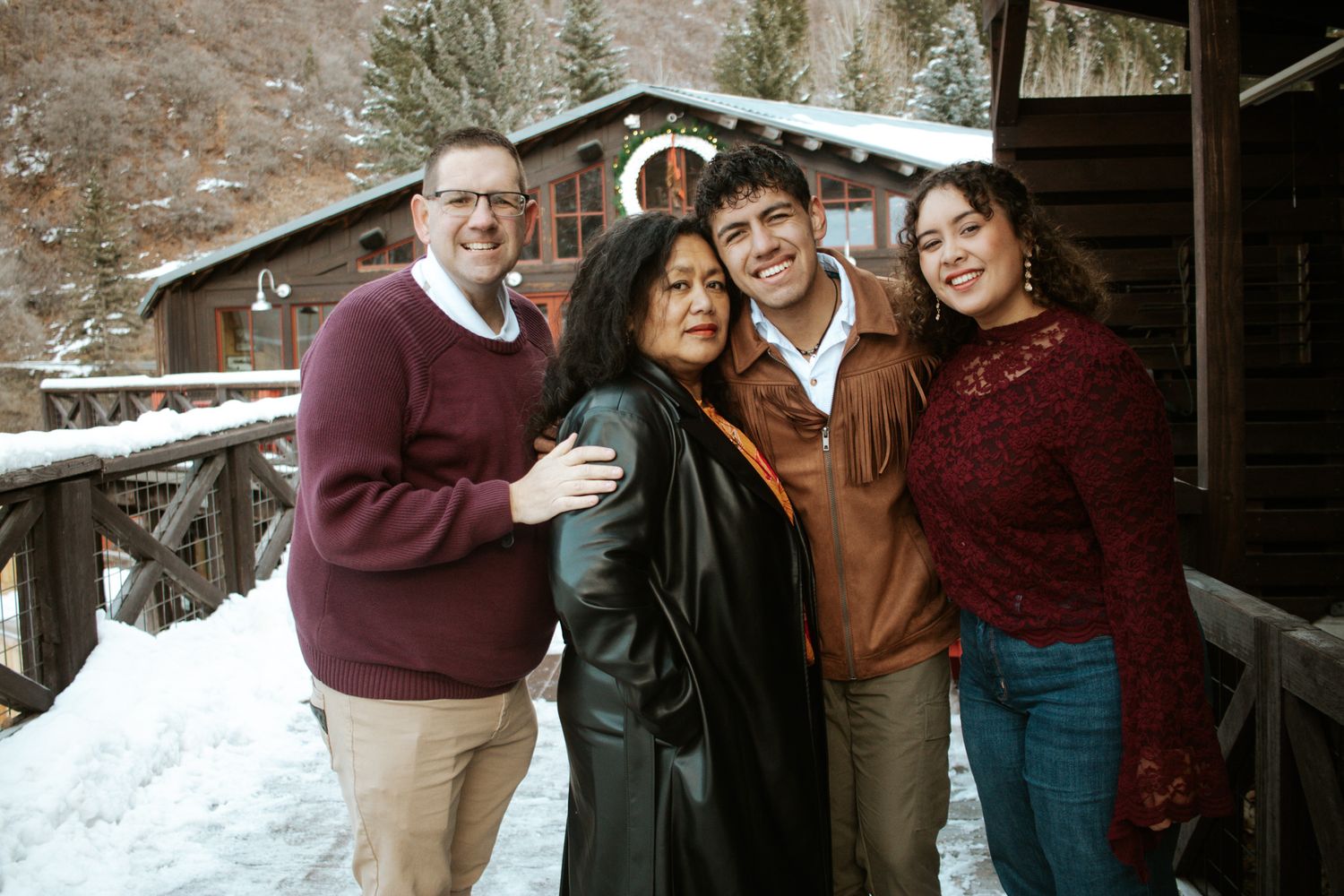 Family of four posing and smiling together outside a snow-covered rustic mountain cabin.
