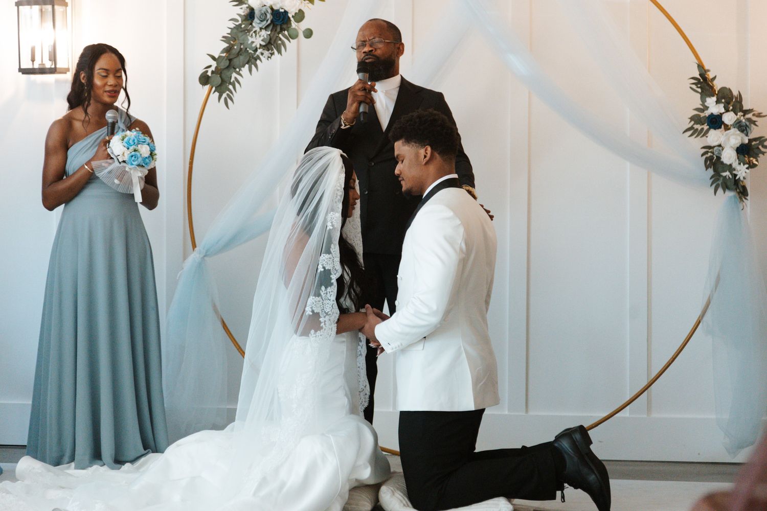 Groom kneeling before bride at elegant wedding ceremony with circular gold arch and dusty blue bridesmaid.