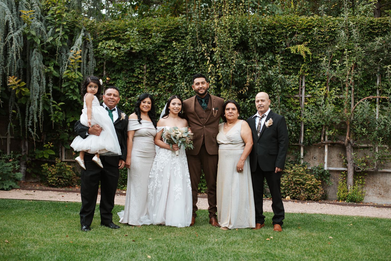 Wedding party poses in white gowns and brown suit with family in lush garden setting.
