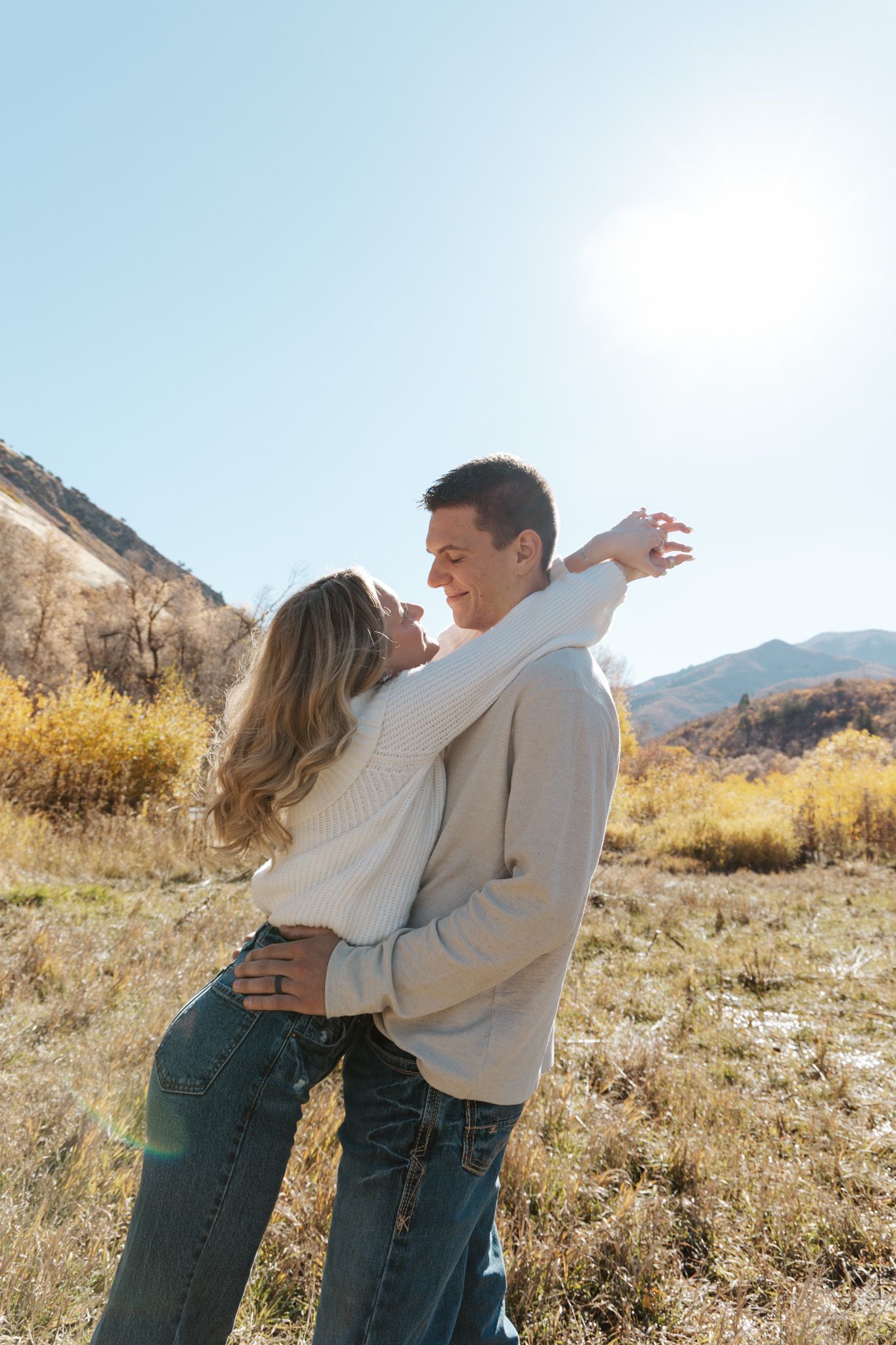 Couple embracing and smiling in a golden autumn mountain field with bright sunlight behind them.