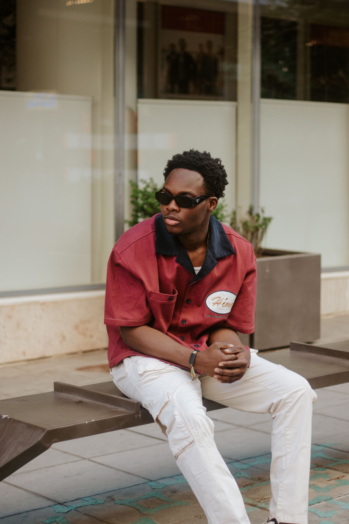 Stylish young Black man in red shirt and white pants sitting on bench outside a shop.