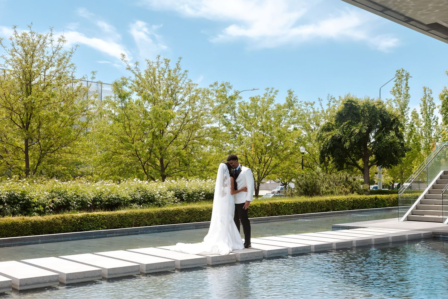Bride and groom share a romantic kiss beside a reflecting pool, surrounded by lush green trees on a sunny wedding day.