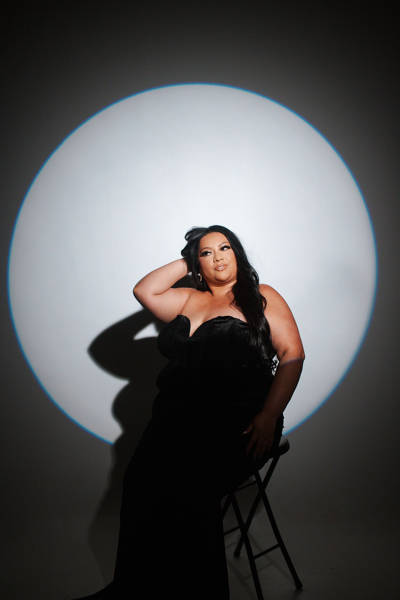 Elegant woman in black strapless gown posing on chair against dramatic spotlight background.