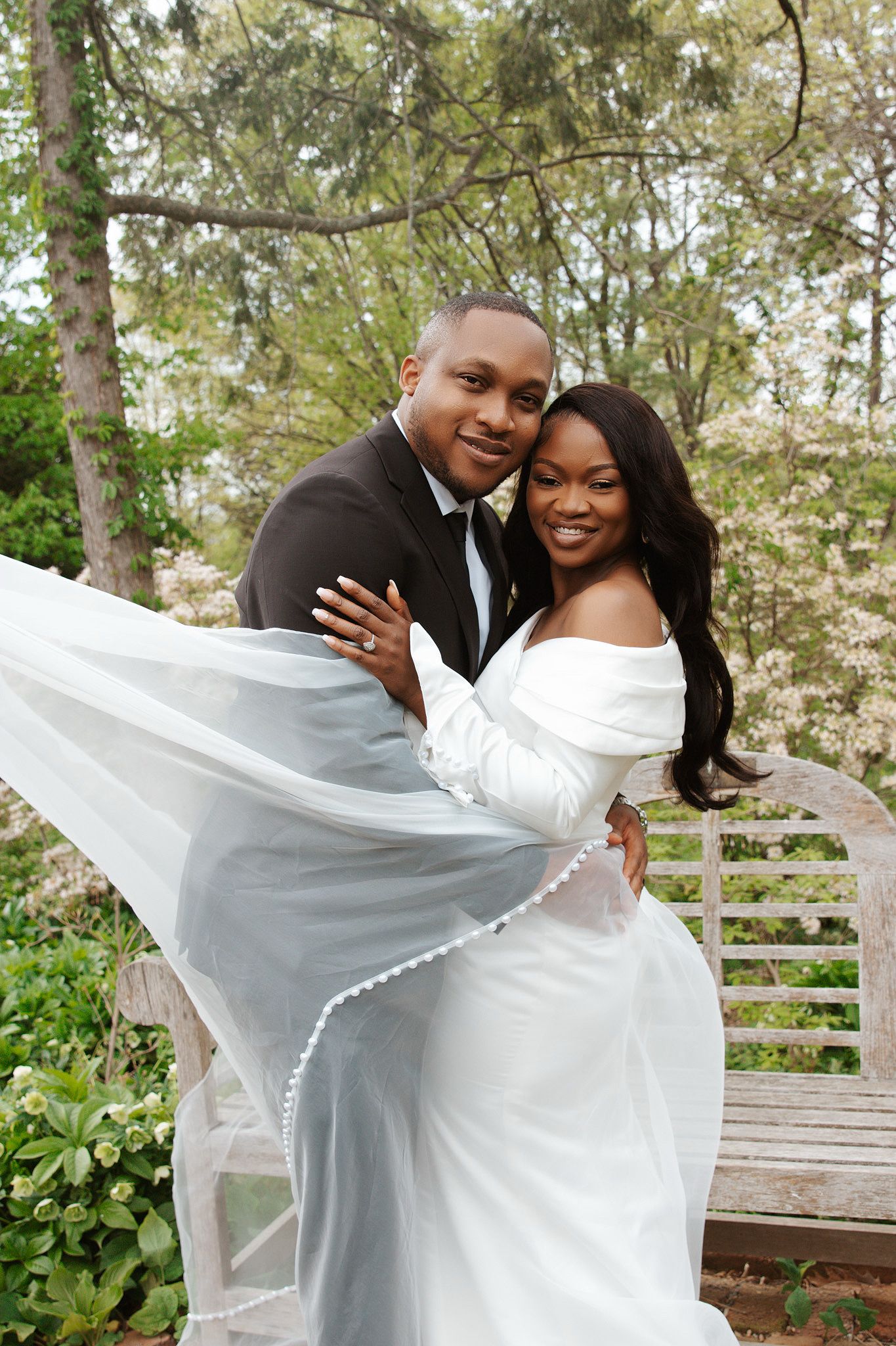 Smiling bride and groom embrace outdoors with flowing veil and blooming trees in background.