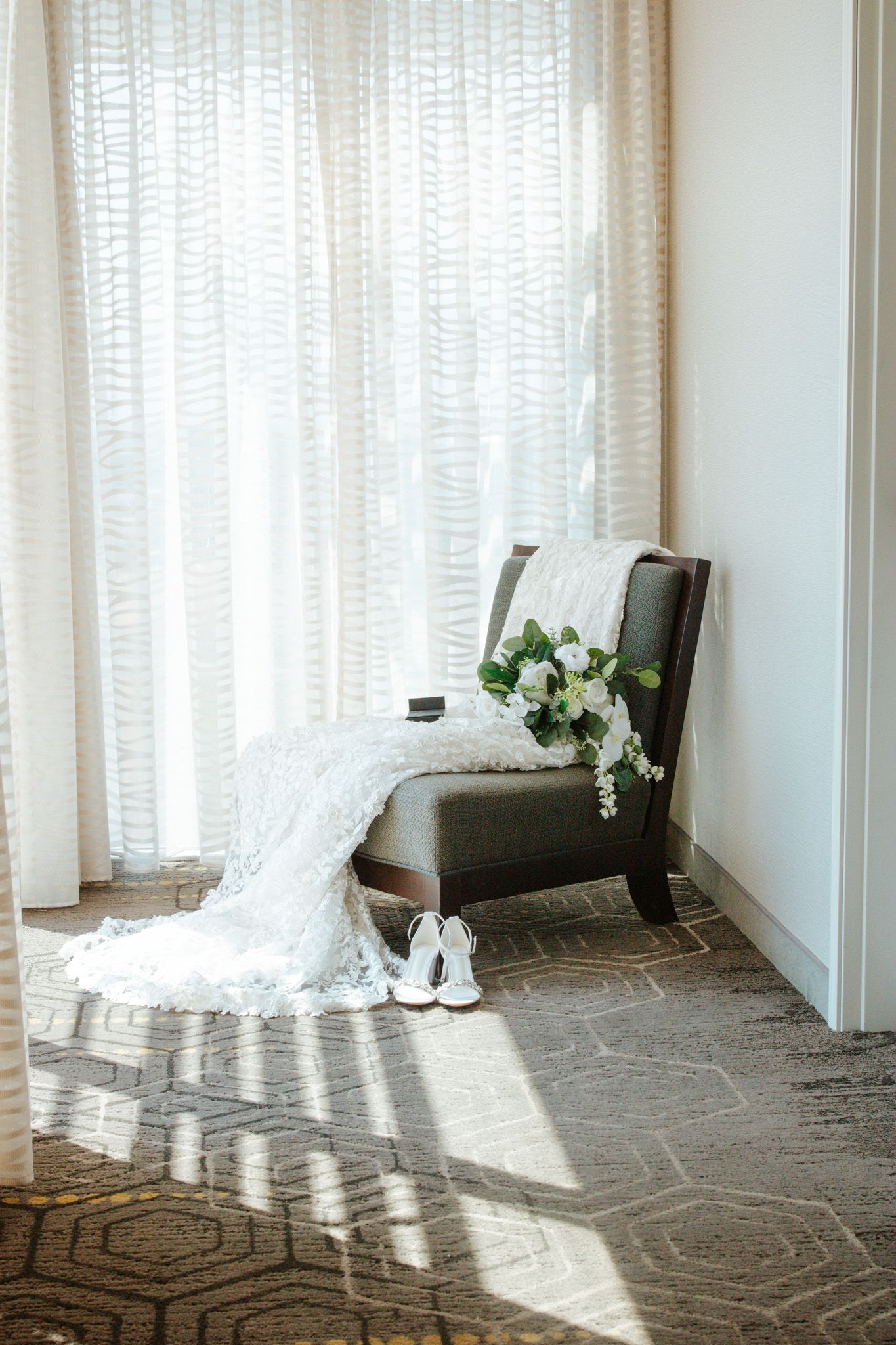 Bridal bouquet of white roses and wedding dress draped over chair with white heels below.
