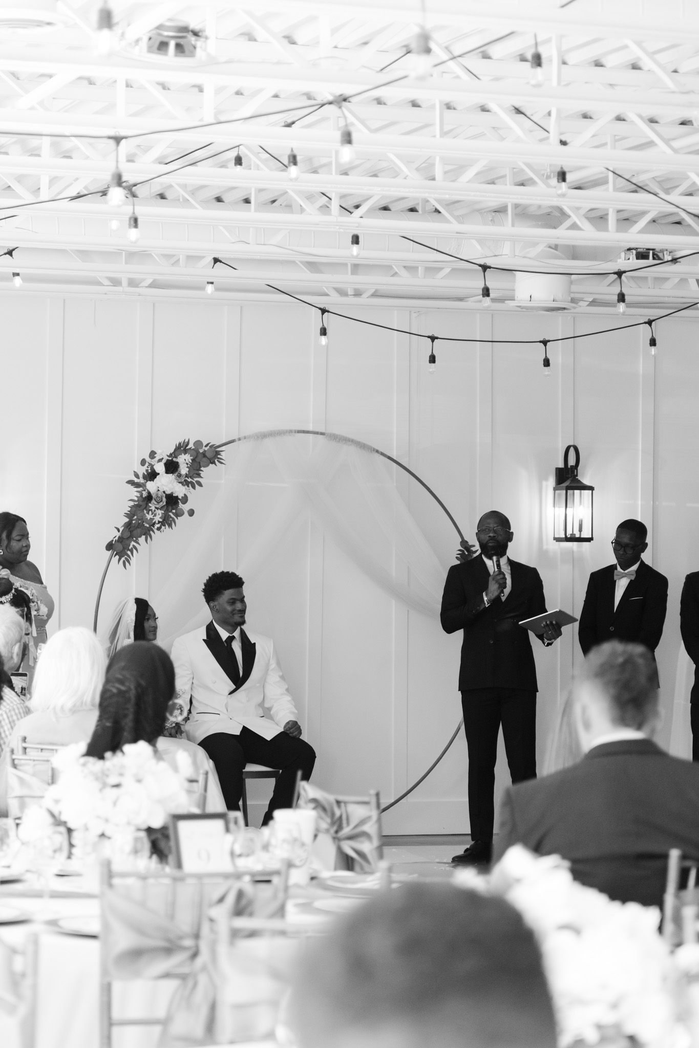 Black and white photo of wedding ceremony with groom seated near circular floral arch as officiant speaks.
