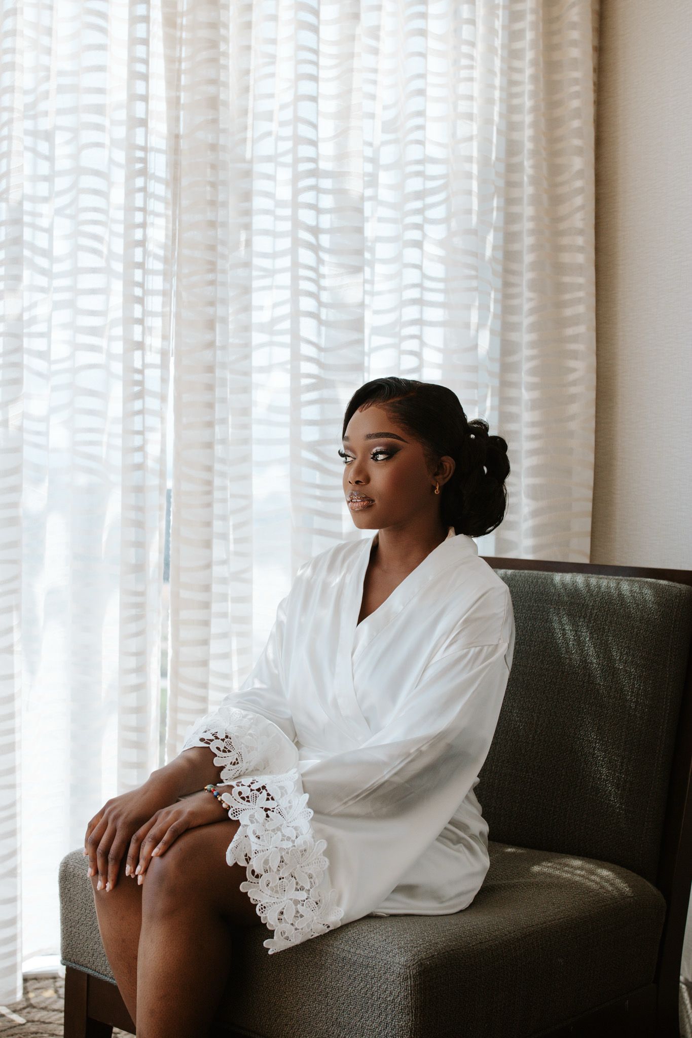 Bride in white lace robe sitting thoughtfully in armchair by sheer curtained window before wedding.