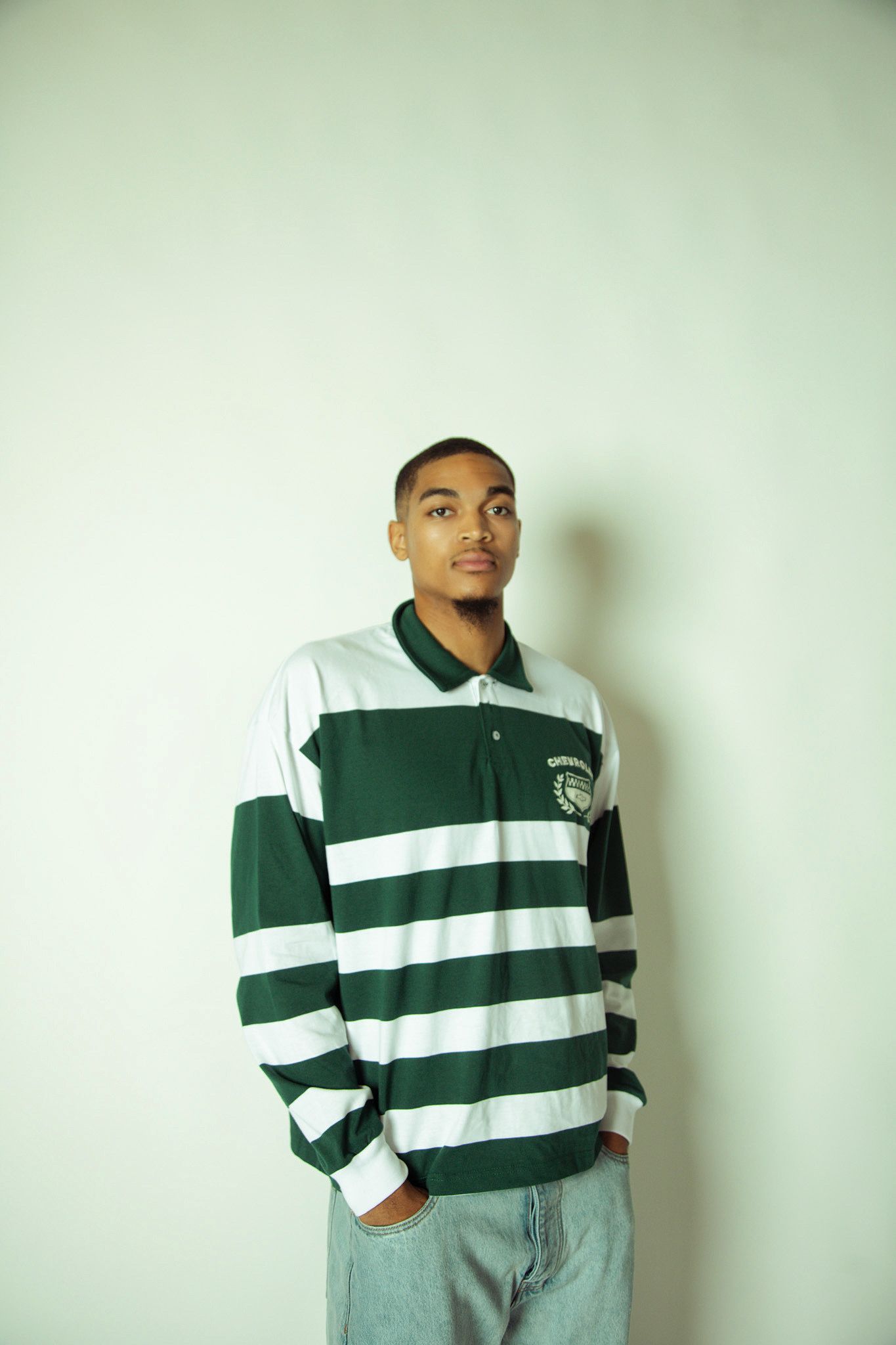 A young Black man in a green and white striped rugby shirt stands against a light gray wall.