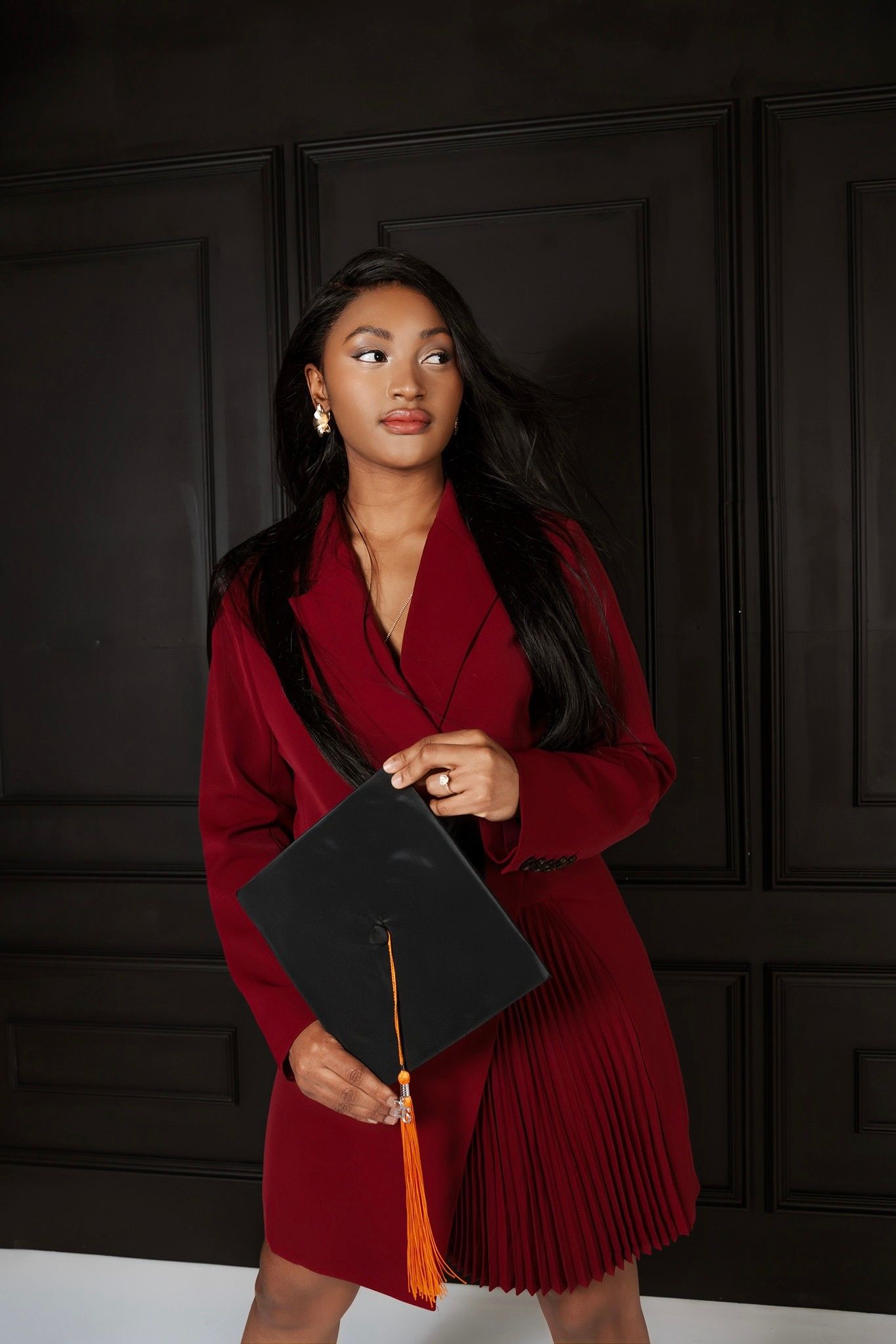 Young woman in red blazer dress holding black graduation cap with orange tassel against dark wall.