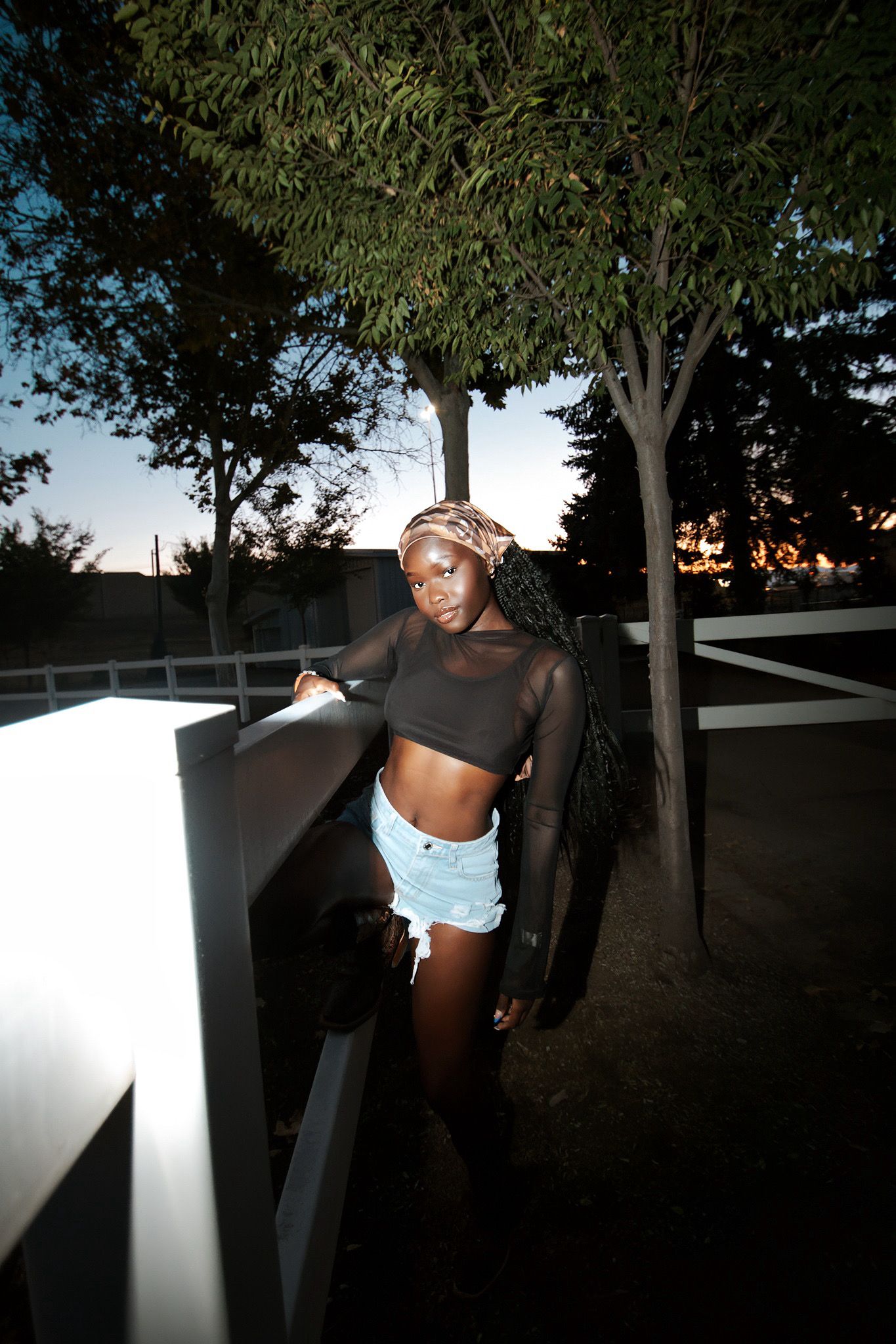 Young Black woman in denim shorts and black mesh crop top leaning on a fence at night.