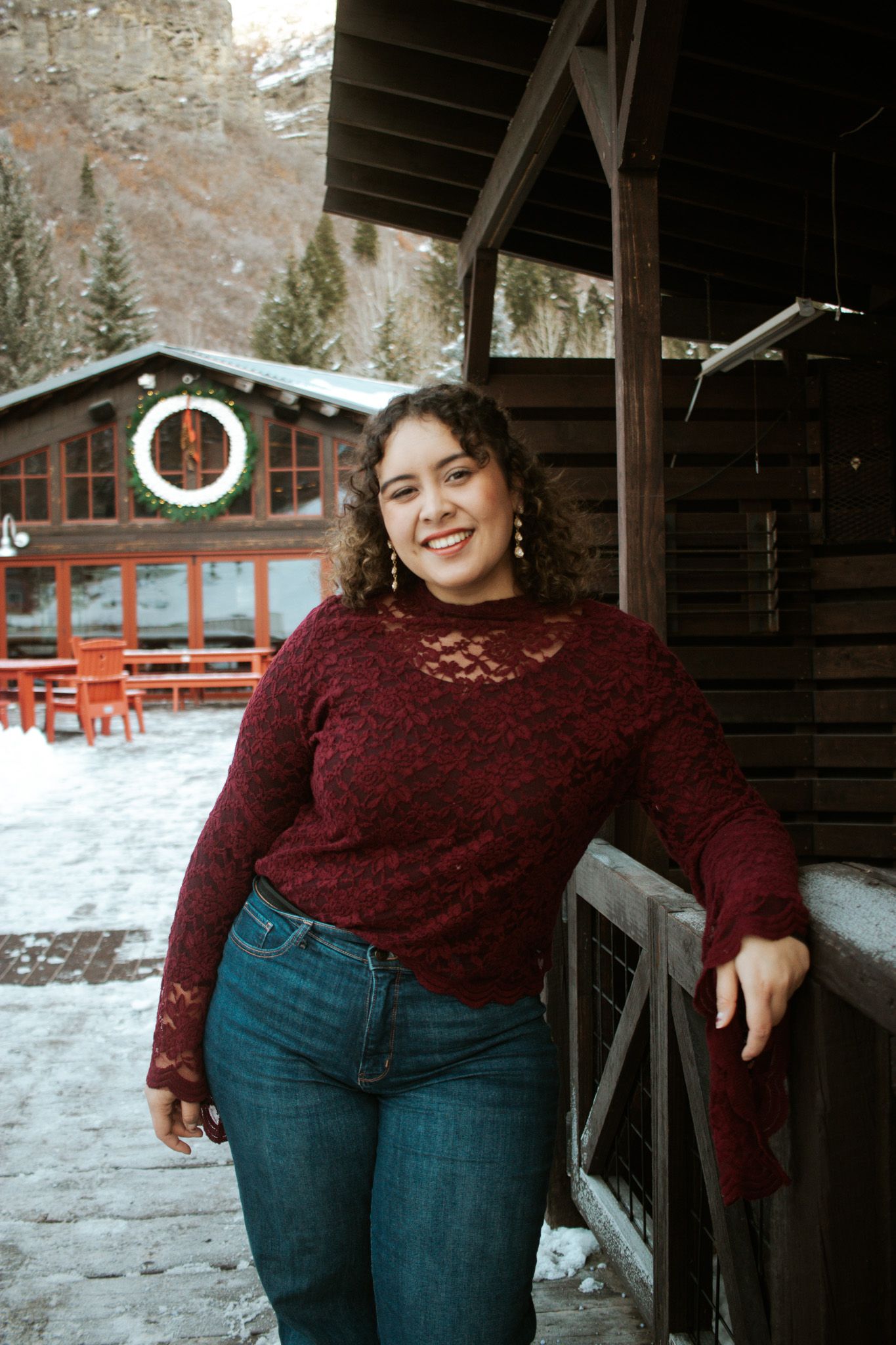 Smiling curly-haired woman in burgundy lace top and jeans posing at snowy mountain lodge.
