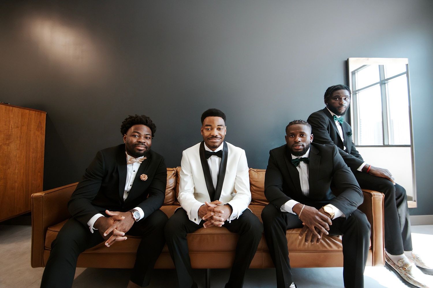 Groom in white tuxedo seated with three groomsmen in black tuxedos on leather sofa indoors.